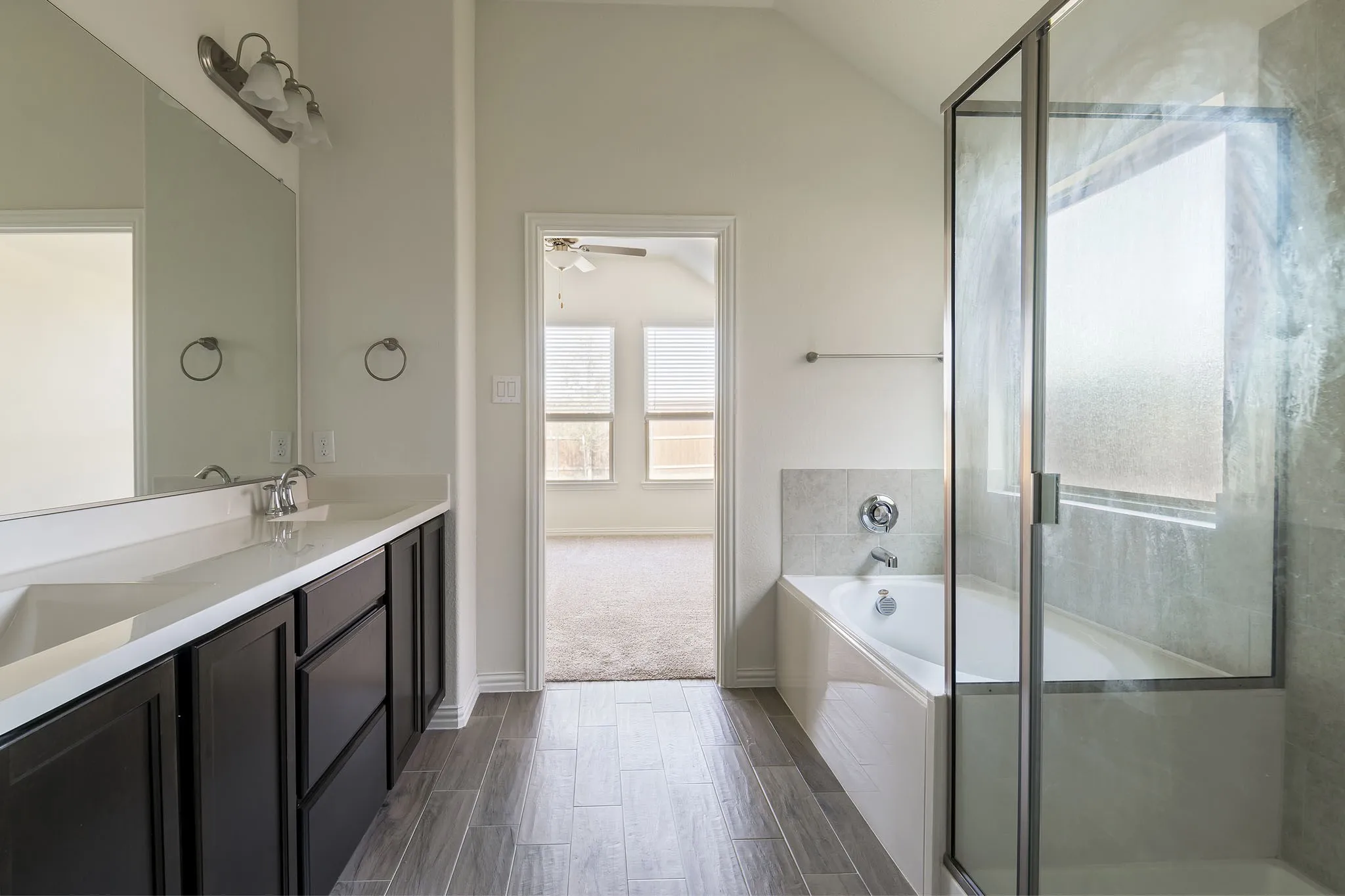 Dual vanity with integrated sinks, framed mirror, wood-finish flooring, built-in bathtub, and glass-enclosed shower