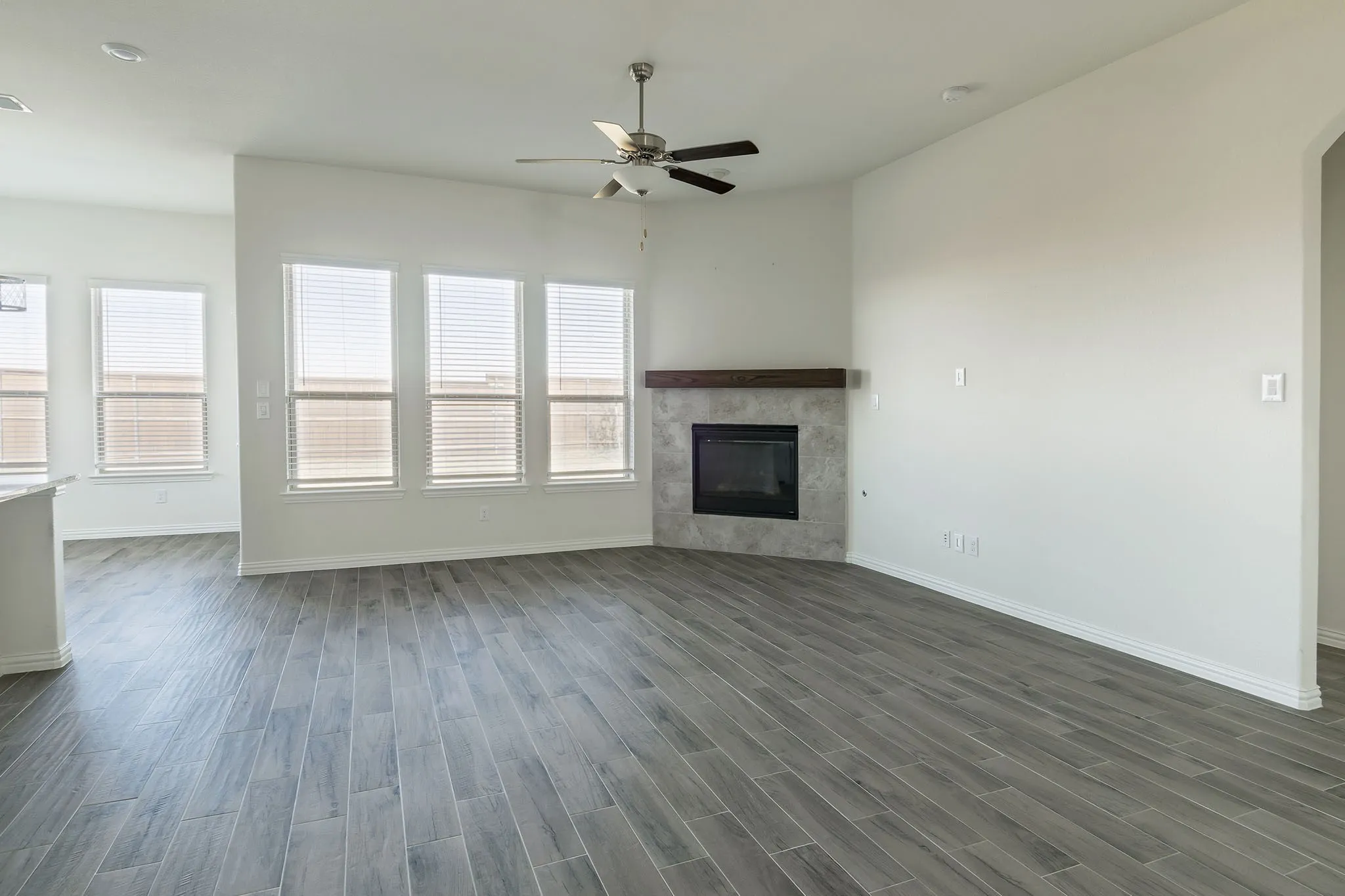 Wood-finish flooring throughout, ceiling fan with integrated lighting, multiple windows providing natural light, tiled fireplace with a wood mantel, and recessed lighting