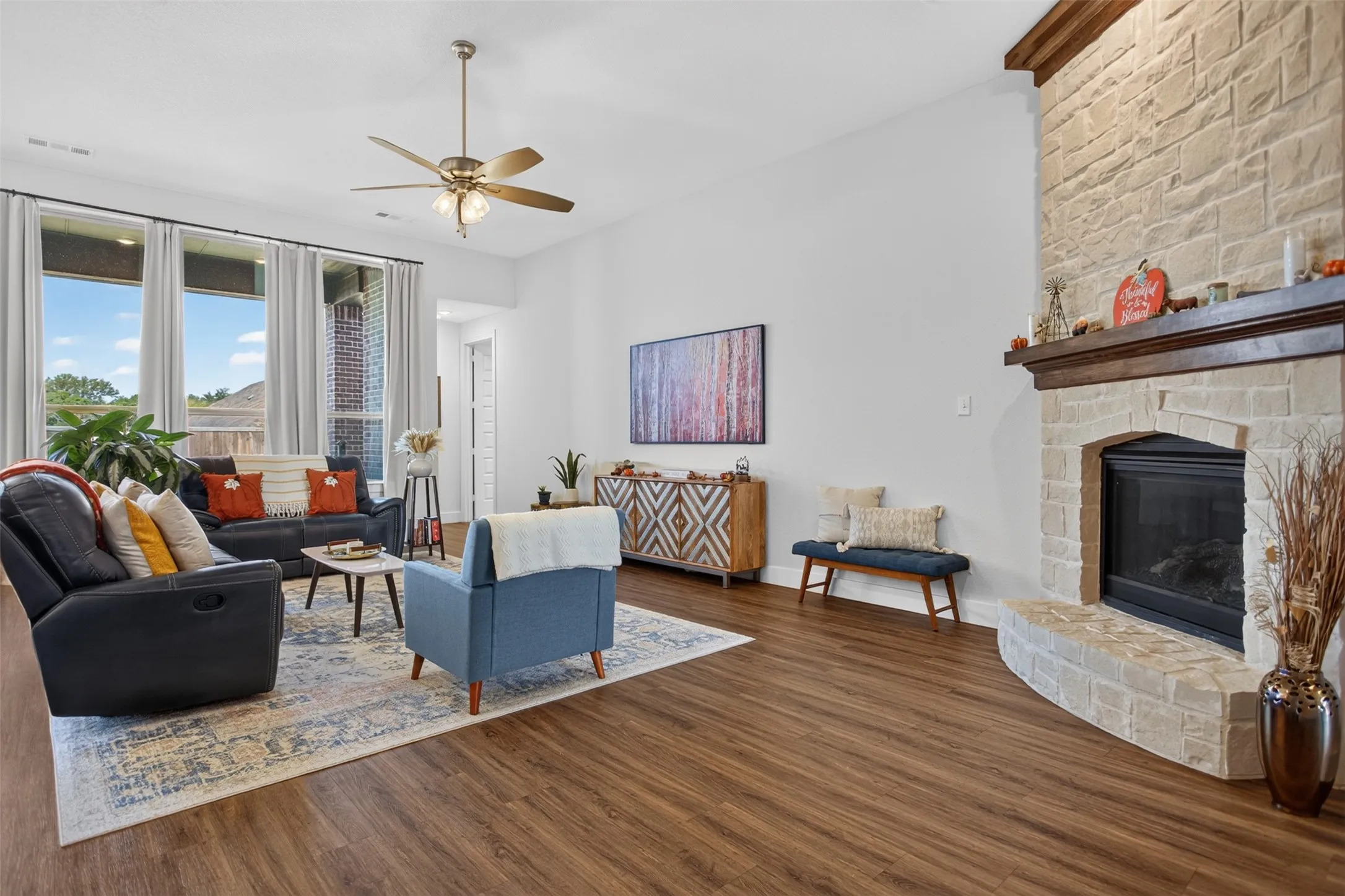 Light-toned stone fireplace with a wood mantle, wood-finish flooring, a ceiling fan with integrated lighting, and multiple large windows