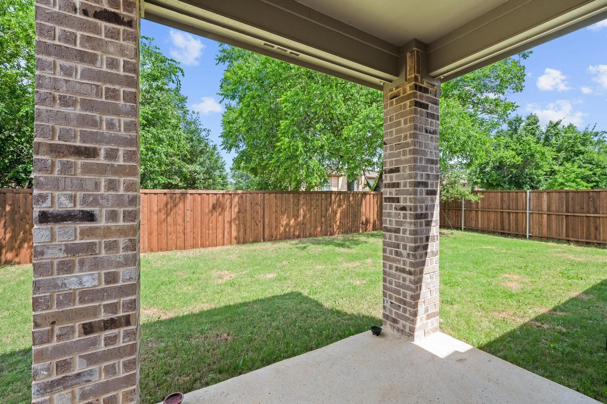 Covered patio with a concrete surface, brick columns, and a fully fenced backyard featuring a grassy lawn and mature trees