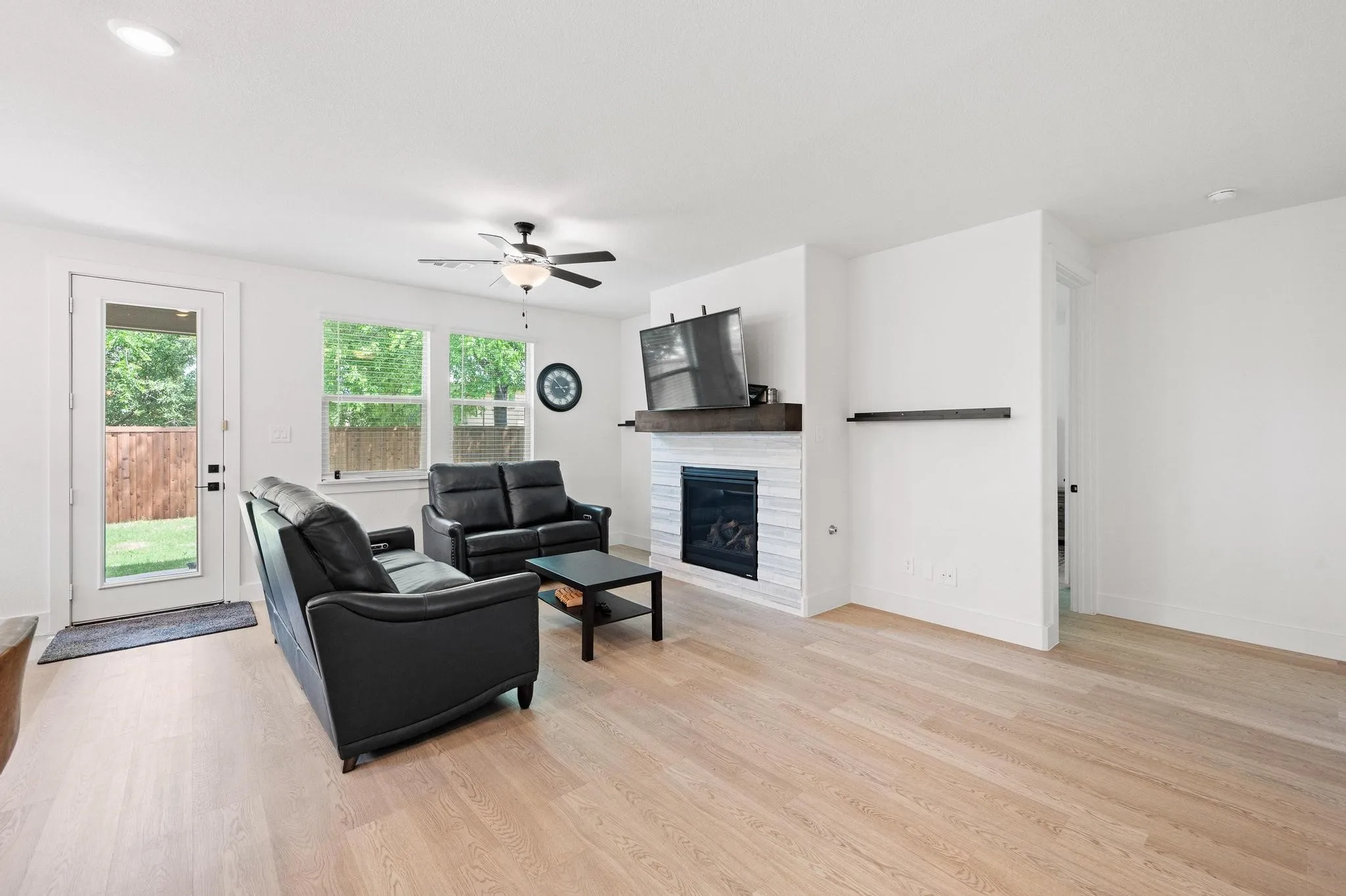Light wood-finish flooring throughout, modern electric fireplace with a wood mantel, glass-paned door to the exterior, and a ceiling fan with integrated lighting