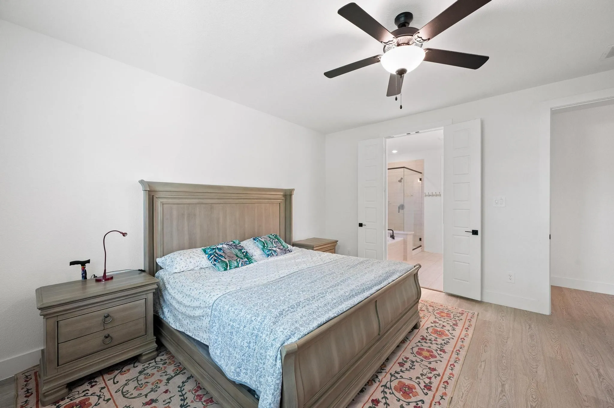 Light wood-finish flooring, ceiling fan with integrated lighting, white walls, and double pocket doors leading to an adjacent bathroom