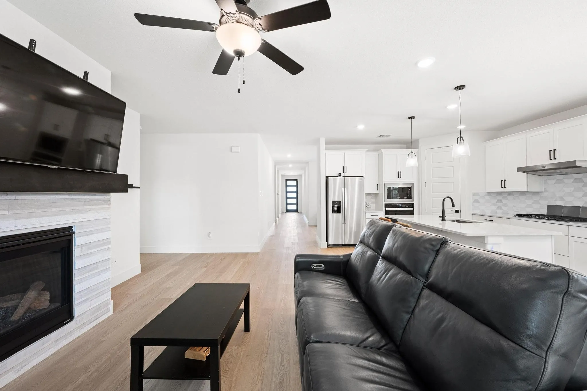 Integrated kitchen and living area featuring wood-finish flooring, a contemporary fireplace with tile surround, white cabinetry, stainless steel appliances, and an island with pendant lighting