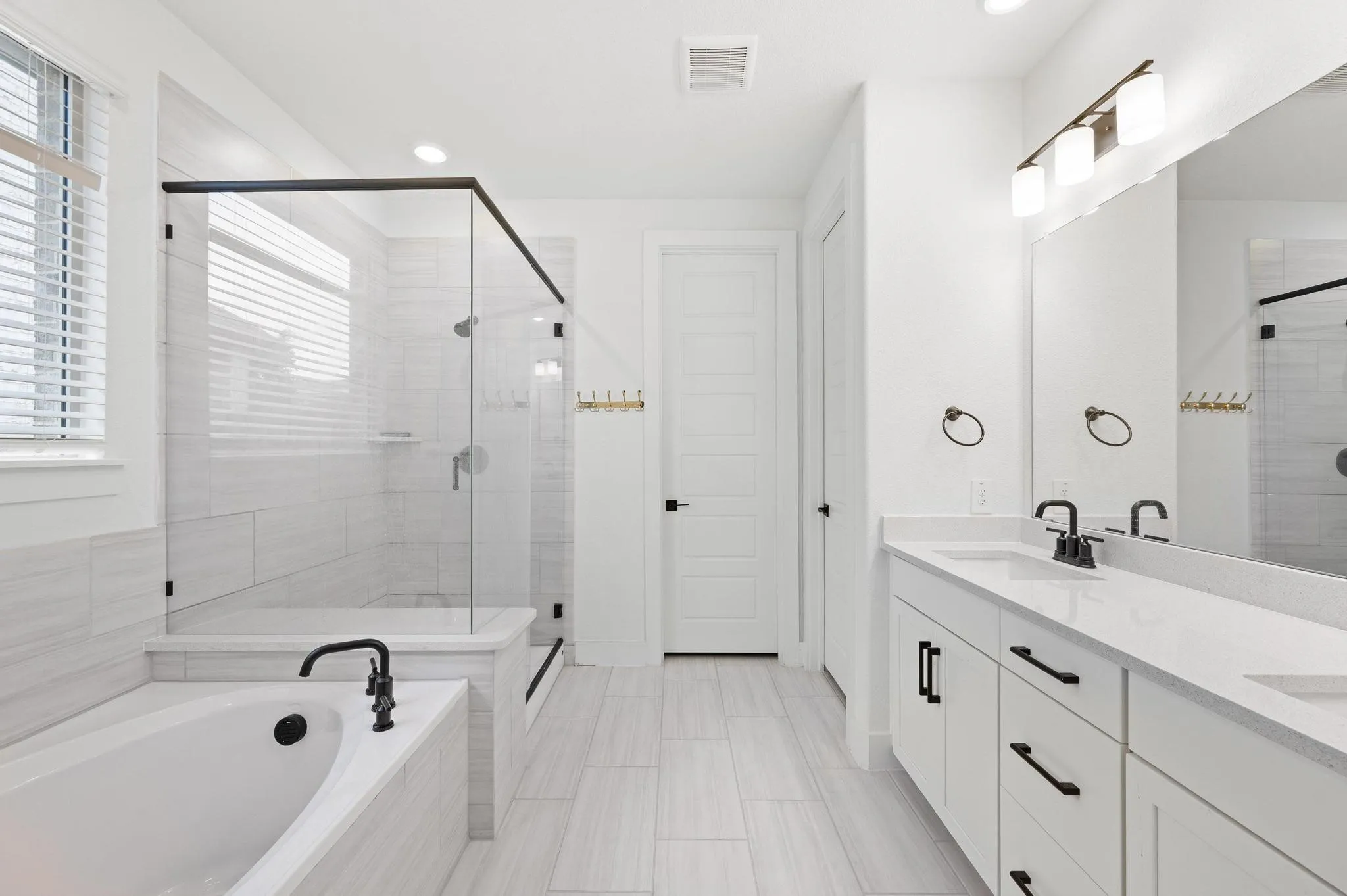 Dual vanity with quartz countertops, a soaking tub with black fixtures, a walk-in shower with clear glass enclosure, white tile flooring, and white paneled doors