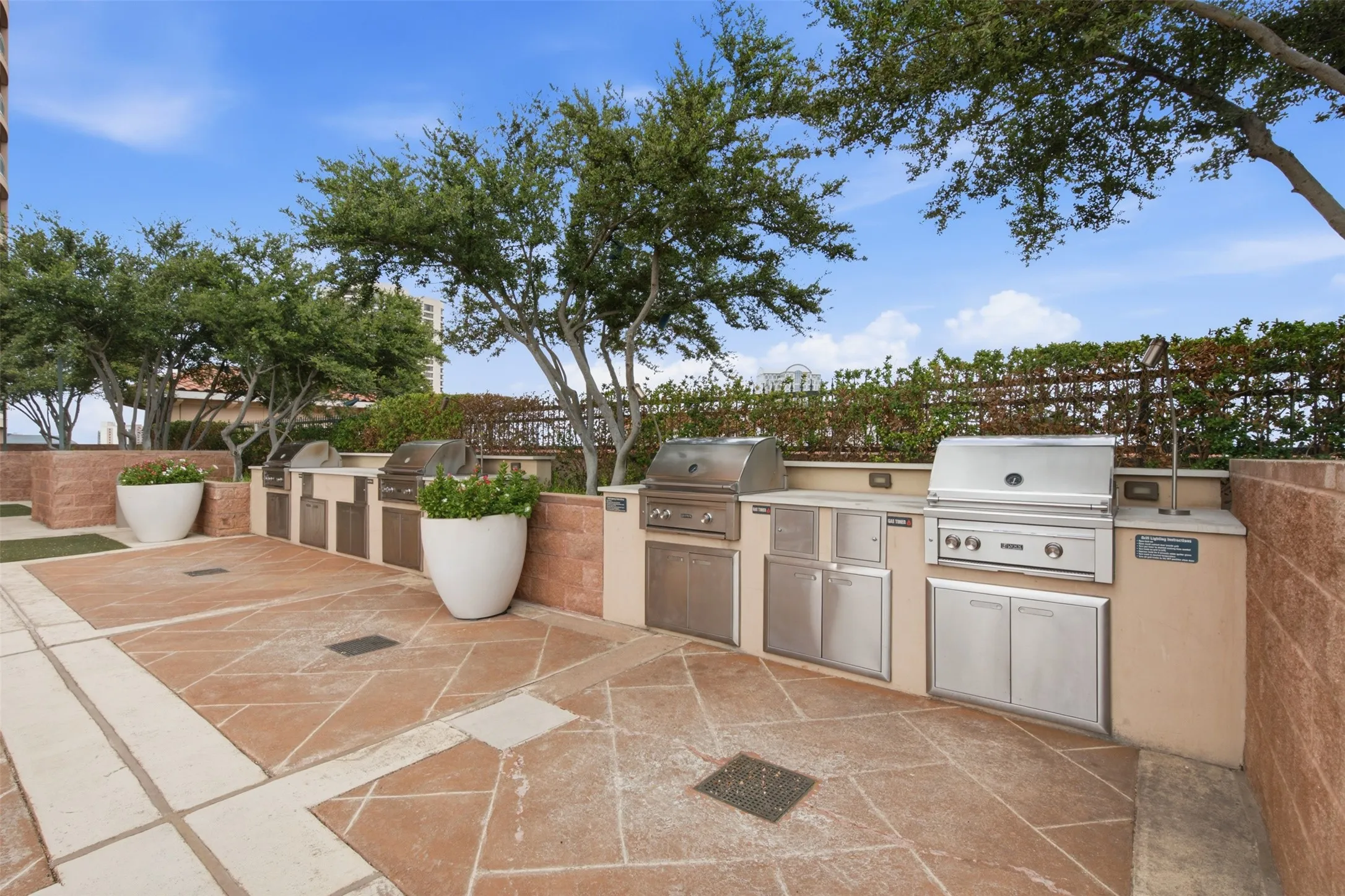 Expansive outdoor patio with stone paver flooring, multiple built-in stainless steel grills, integrated cabinetry, and lush perimeter landscaping