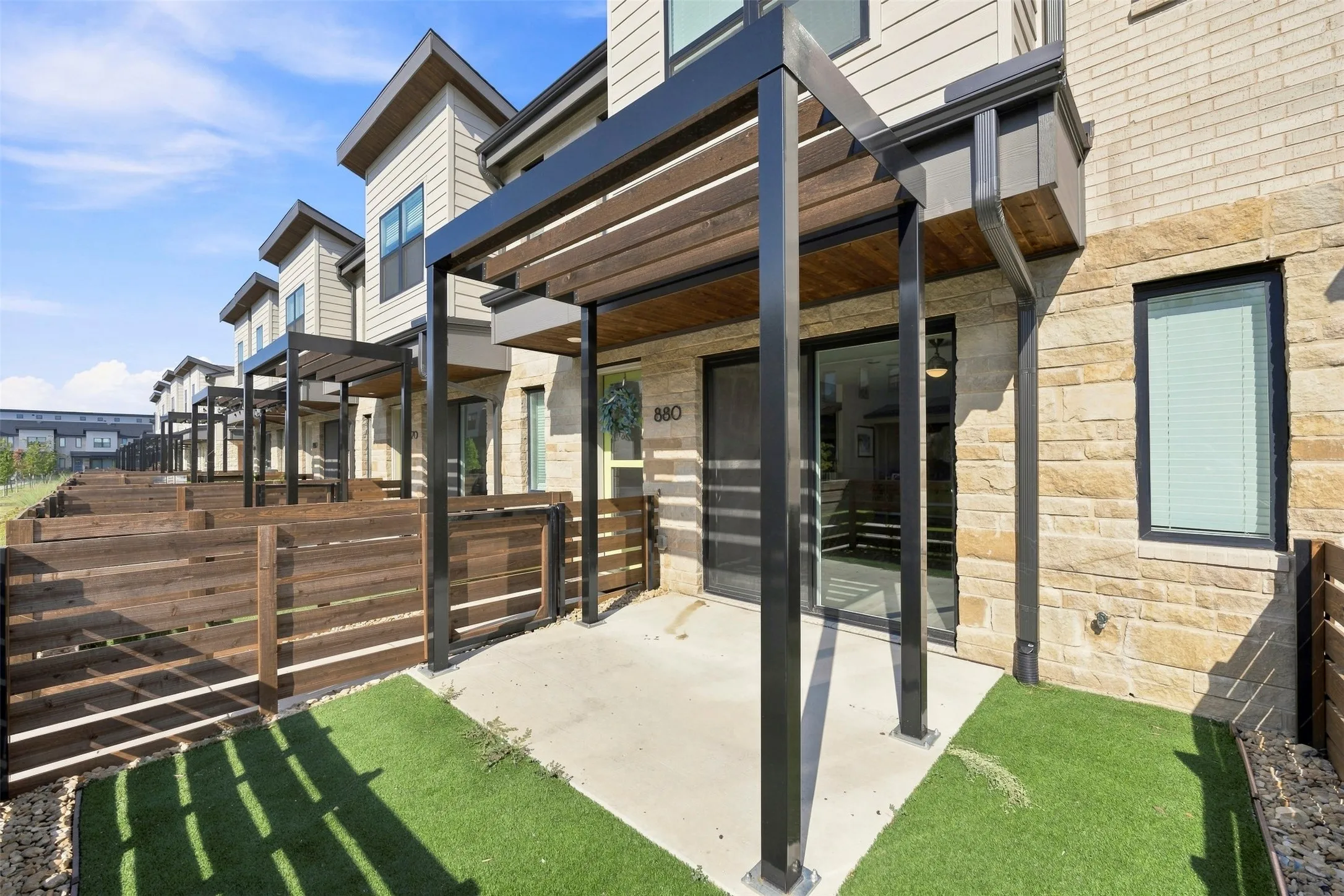 Contemporary townhome exterior featuring stone and siding, a covered patio with a wood slat ceiling, black metal supports, a sliding glass door, and faux turf landscaping