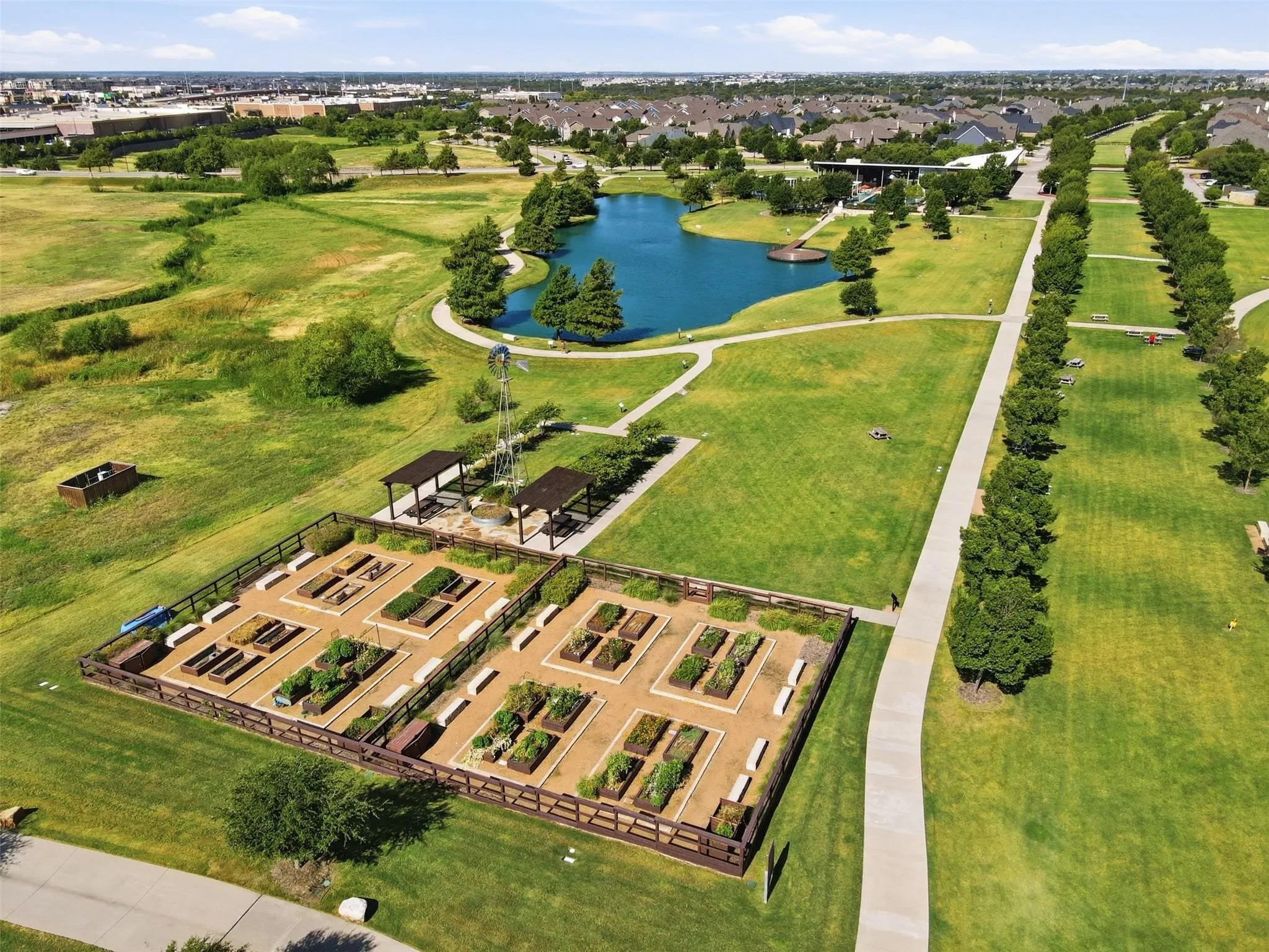 Community garden plots with raised beds, adjacent to walking paths, a pond, and shaded pergola areas