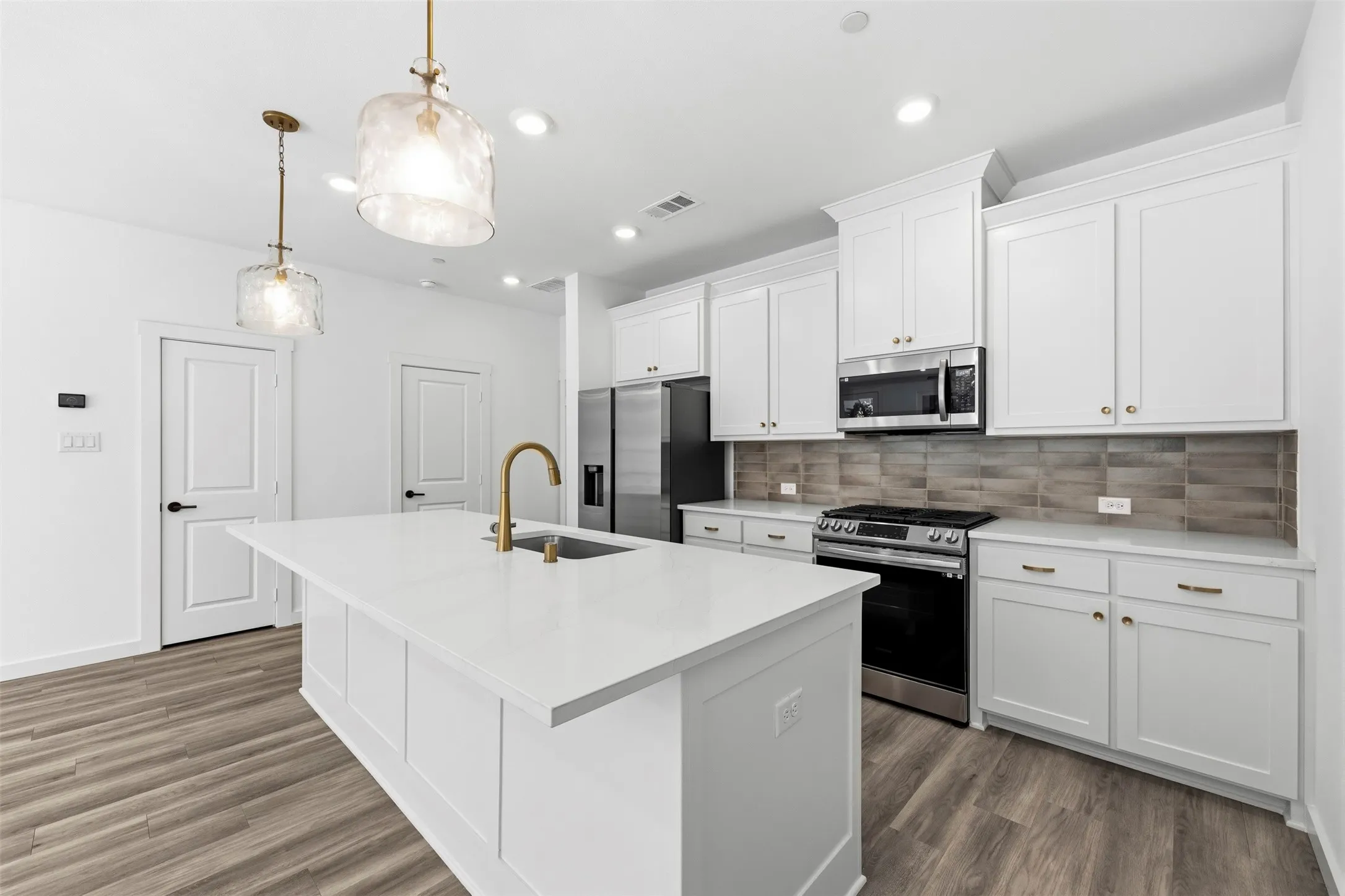 Modern kitchen featuring white shaker-style cabinetry, a large waterfall-edge island with seating, stainless steel appliances including a gas range and built-in microwave, quartz countertops, a subway tile backsplash, brass hardware accents, pendant lighting, and wood-finish flooring throughout the open-concept space