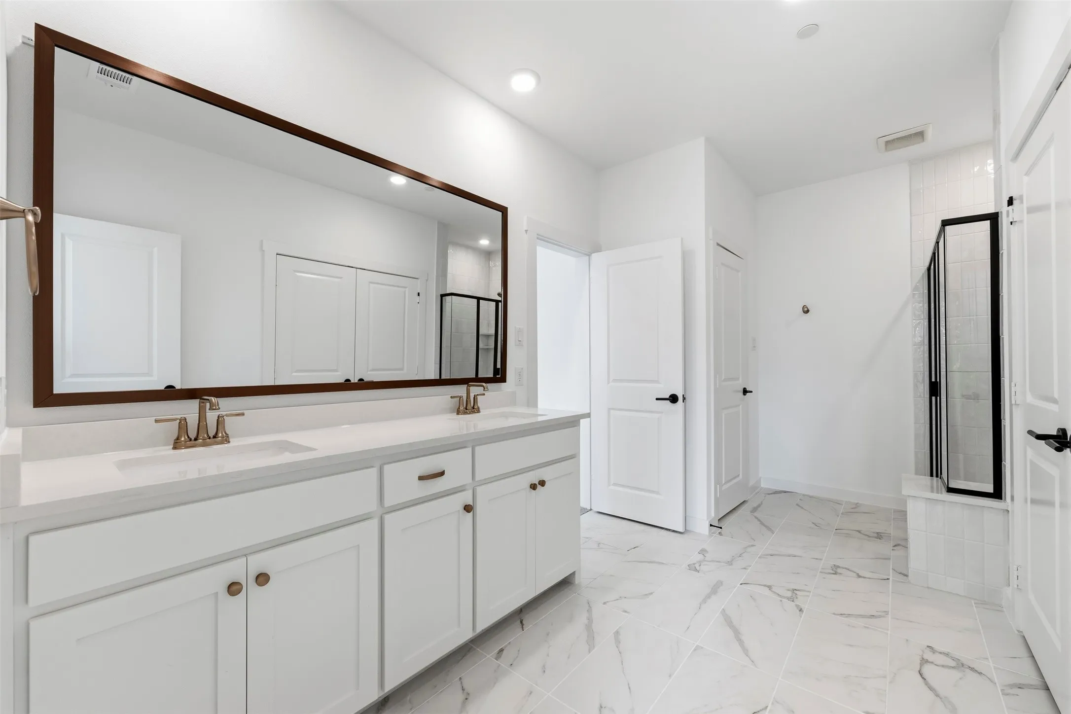 Dual vanity with quartz countertop, wood-finish framed mirror, white cabinetry, and brushed bronze fixtures