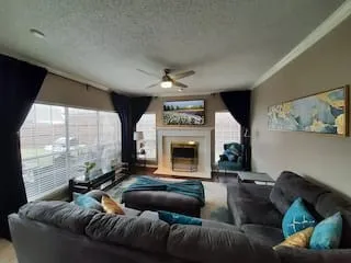 Spacious living area featuring large windows, a ceiling fan with integrated lighting, crown molding, and a built-in fireplace with a mantel