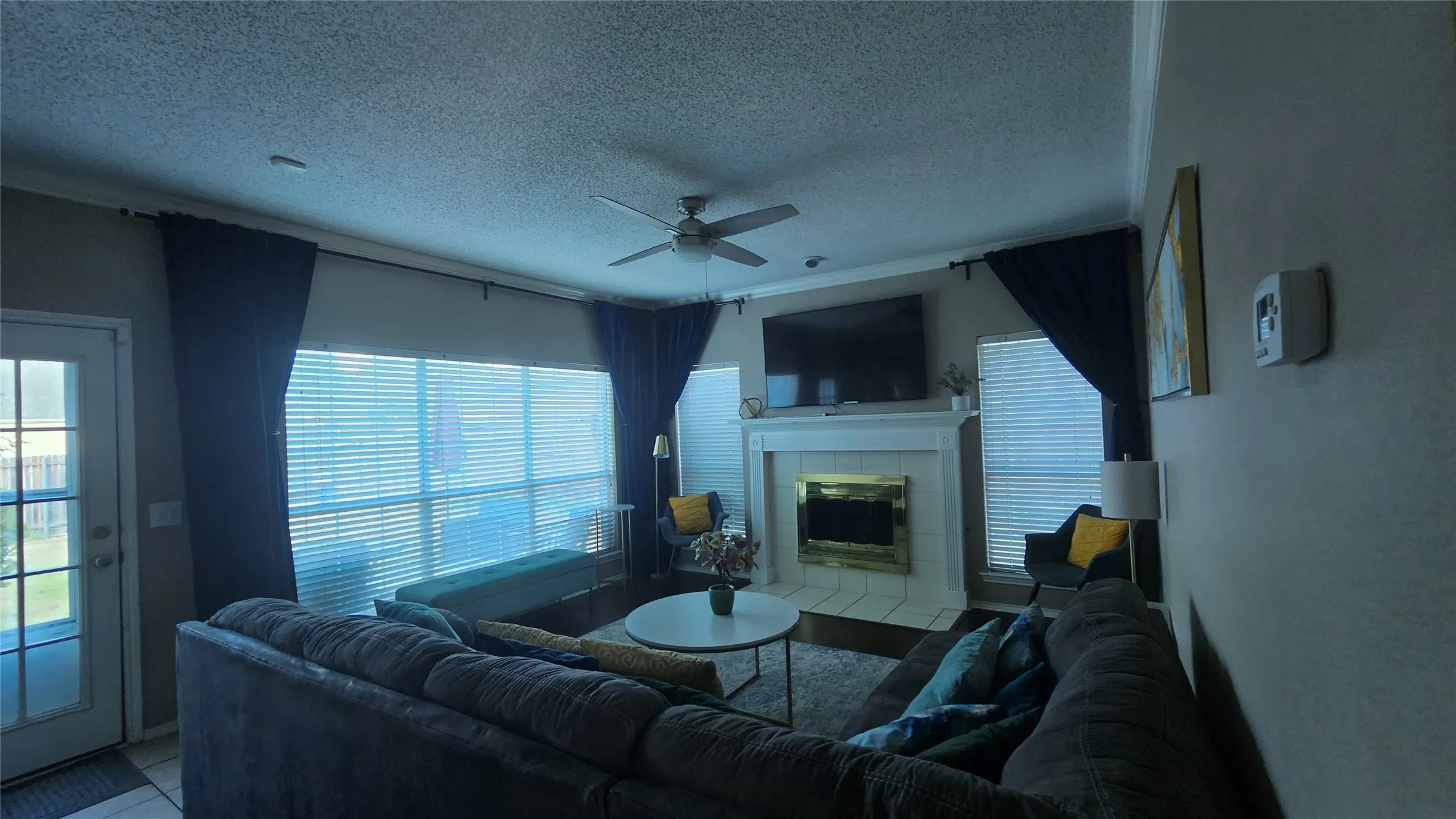 Living area with a ceiling fan, a fireplace with a white mantel, and a large window with blinds