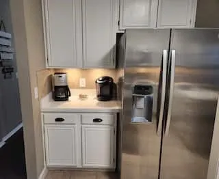 Stainless steel side-by-side refrigerator, white upper and lower cabinetry, light-toned countertop, and under-cabinet lighting