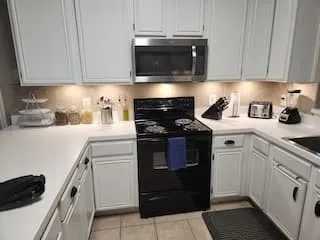 White cabinetry with black hardware, white countertops, built-in stainless steel microwave, black range, and tiled flooring