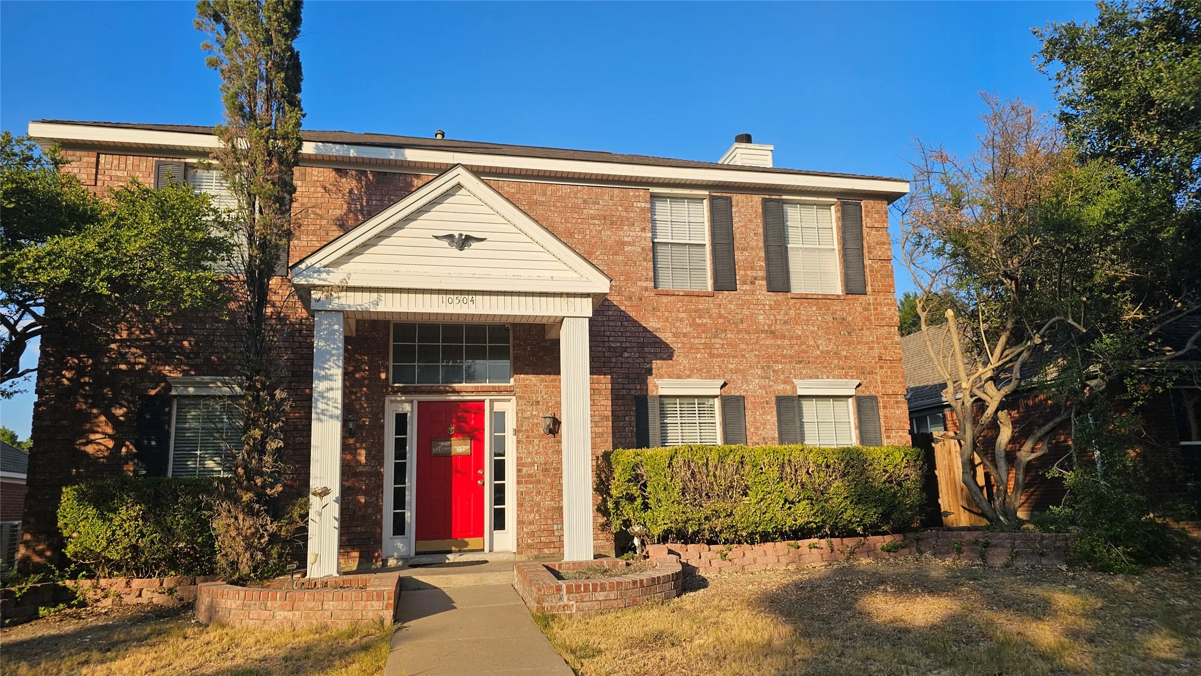 Brick facade, white columns, prominent red entry door, symmetrical window placement, integrated landscaping