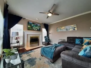 Living area featuring a fireplace with a white mantel, ceiling fan with integrated lighting, wood-finish flooring, and multiple windows with blinds