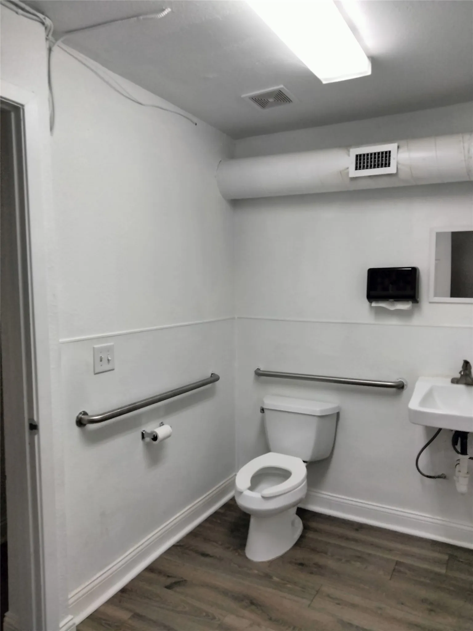 Restroom featuring wood-finish flooring, white wainscoting, wall-mounted sink, and grab bars