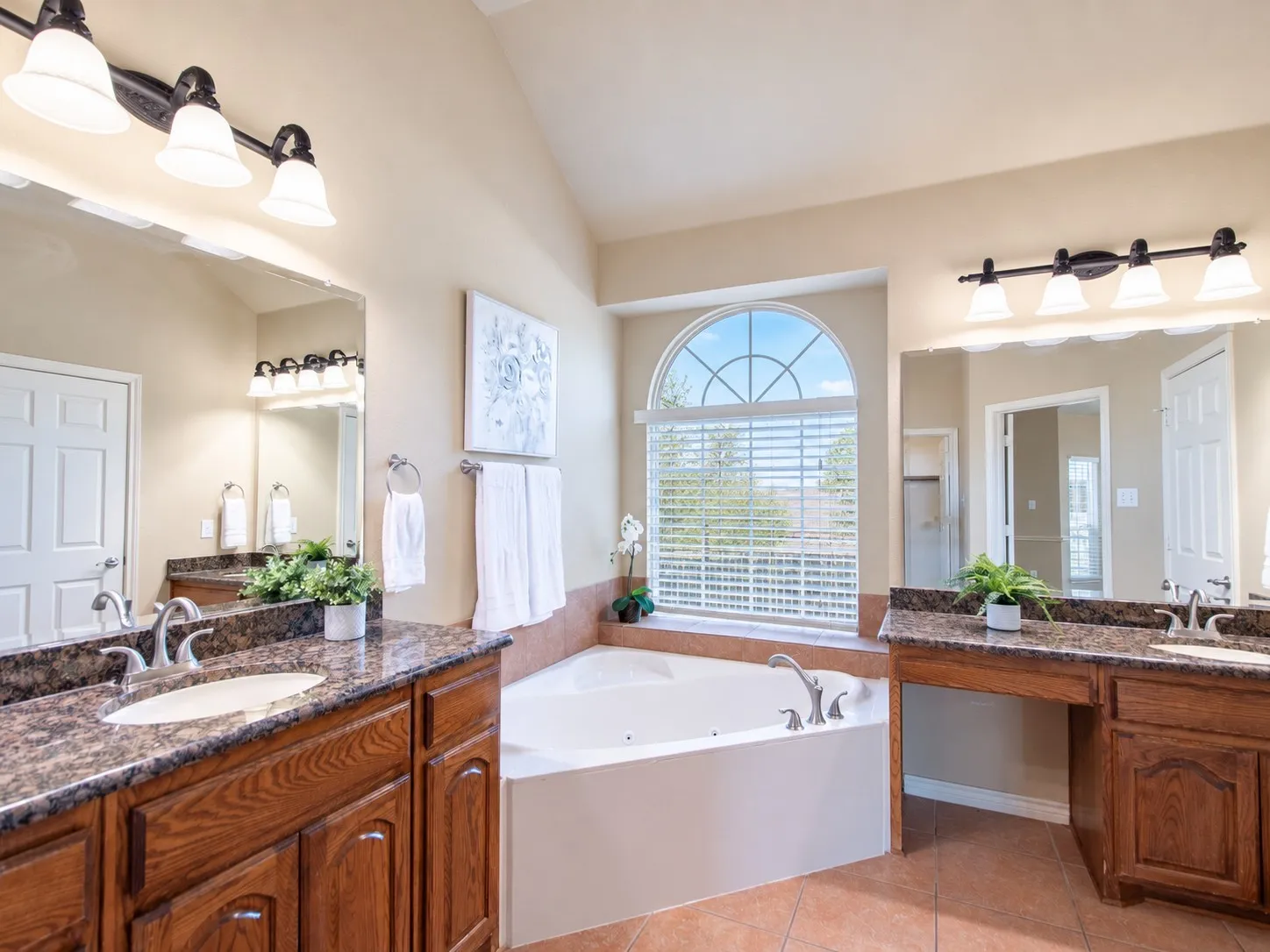 Vaulted ceilings, dual vanities with granite-style countertops, a built-in jetted tub, and an arched window with blinds
