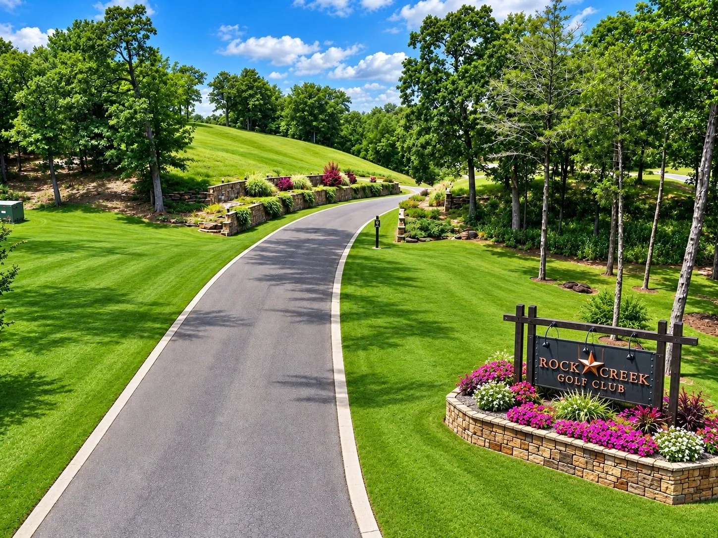 Paved driveway bordered by manicured lawns, stone-faced retaining walls with mature landscaping, and a prominent entry sign