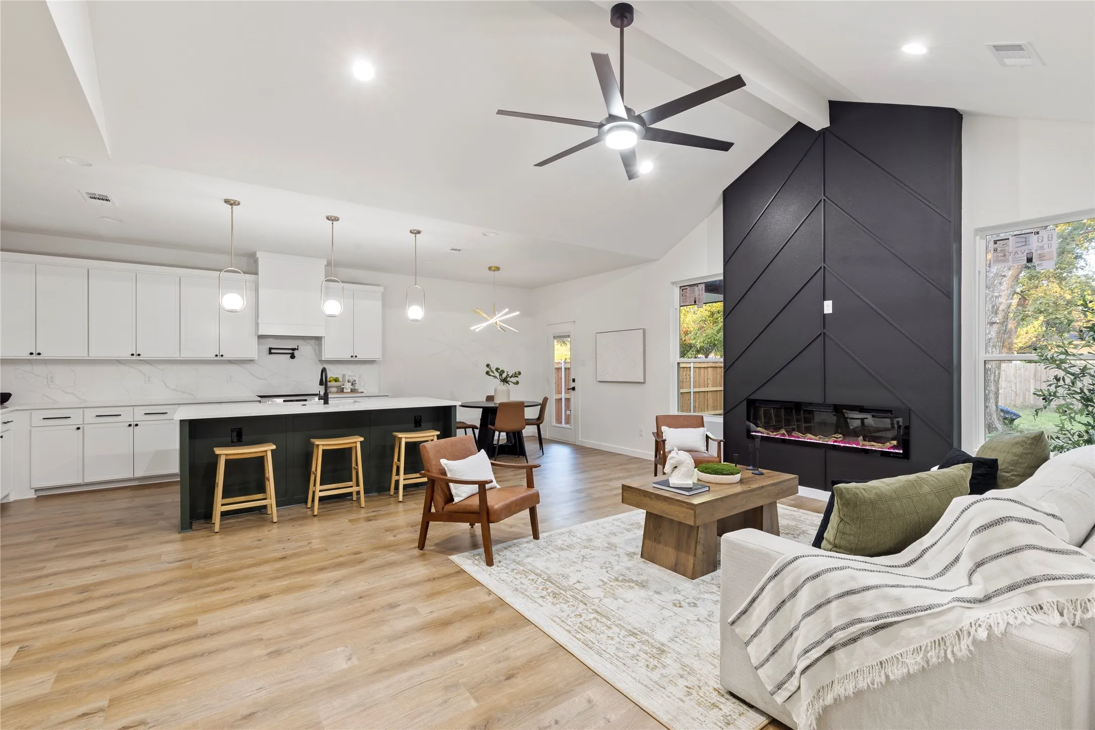 Vaulted ceiling, wood-finish flooring, white cabinetry, kitchen island with seating, and a modern fireplace with a black accent wall