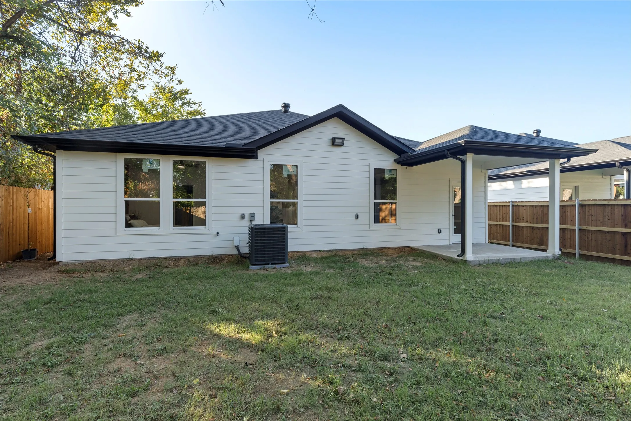 White horizontal siding with a dark roof and gutters, featuring multiple windows, a covered patio with white columns, and a wood privacy fence