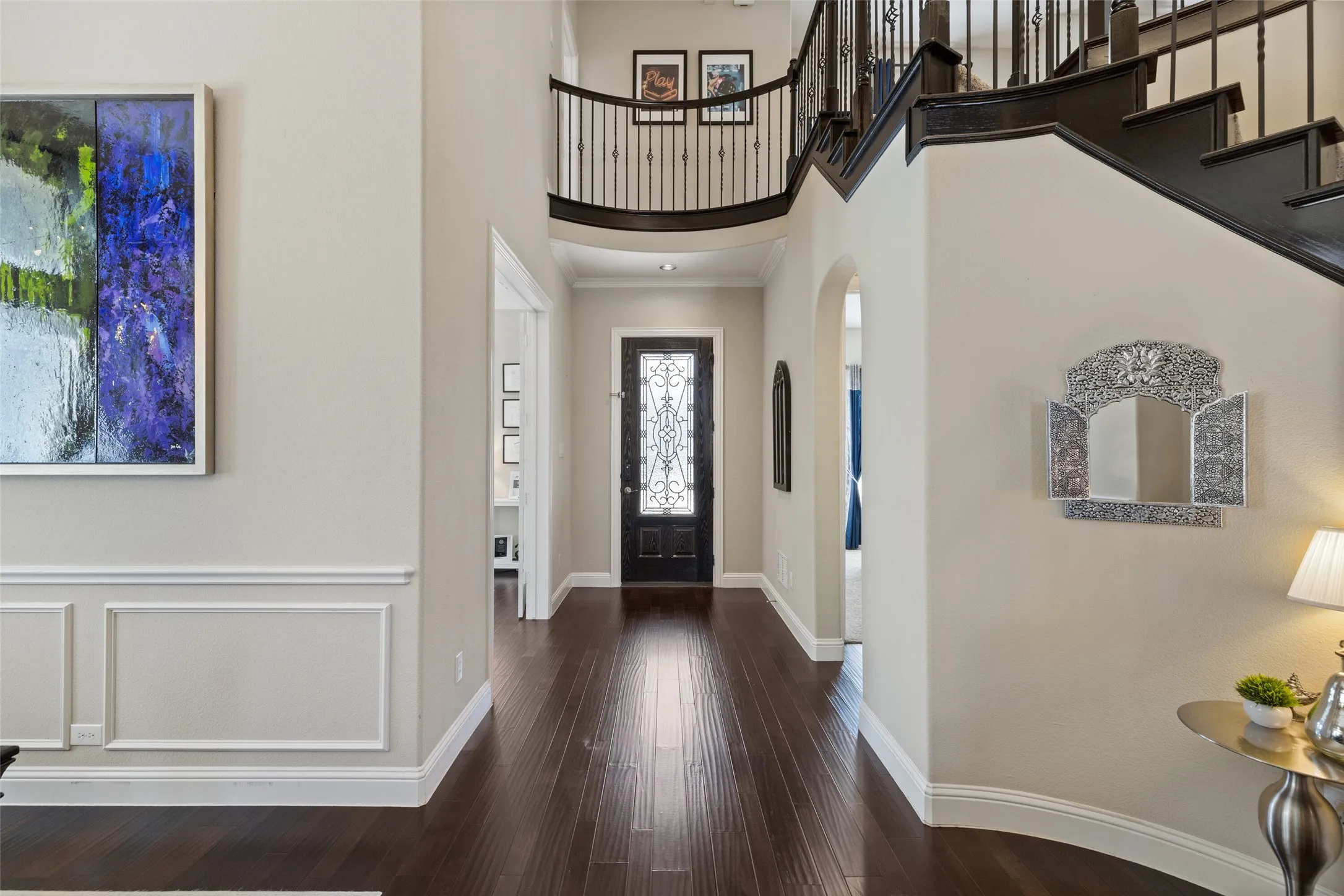 Two-story foyer featuring dark wood-finish flooring, a grand staircase with wrought iron balusters, a decorative entry door with glass inserts, and wainscoting details