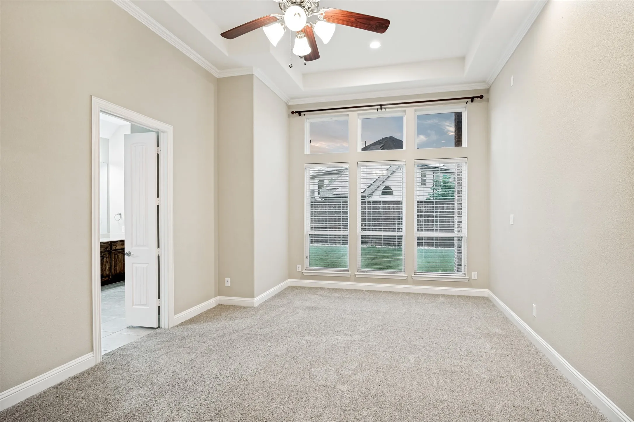 Tray ceiling with crown molding, ceiling fan with light fixture, large multi-pane windows, light-colored carpet, and an open doorway