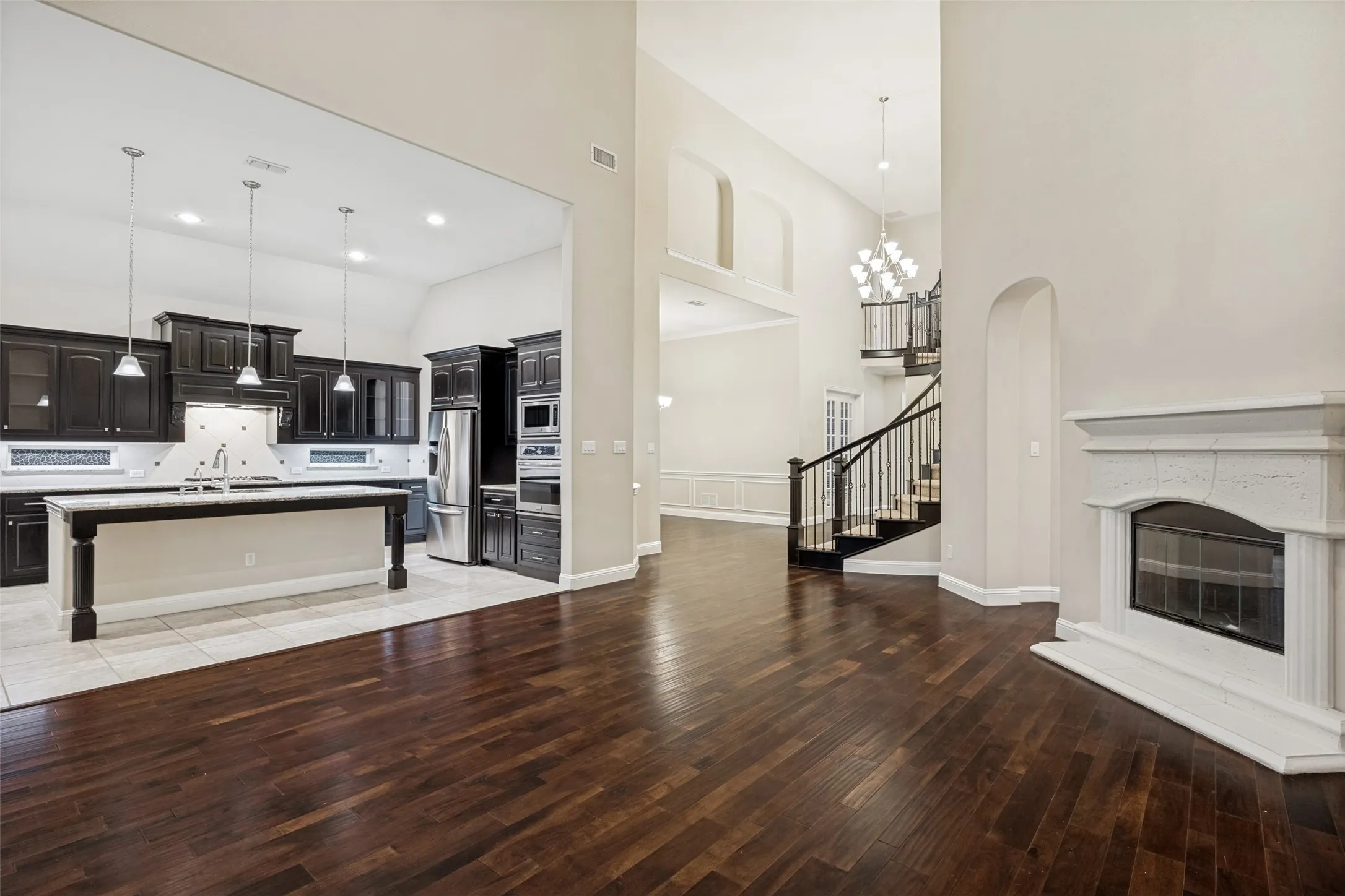 Hardwood-finish flooring, a white stone fireplace, a dark wood staircase with wrought iron balusters, a kitchen island with pendant lighting, and dark wood kitchen cabinetry
