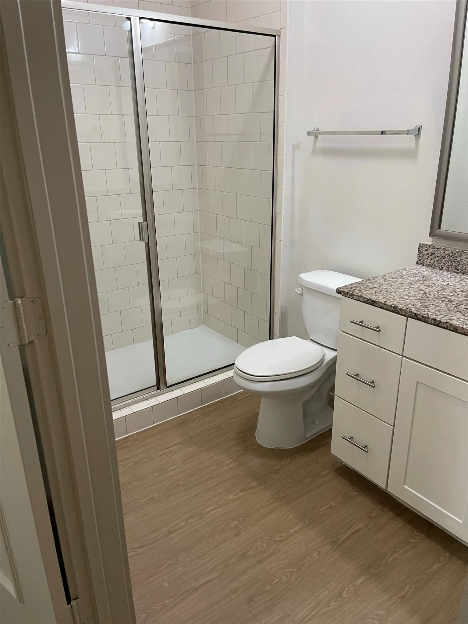 Walk-in shower with clear glass enclosure and white subway tile surround, wood-finish flooring, white vanity with granite-style countertop, and silver-tone hardware