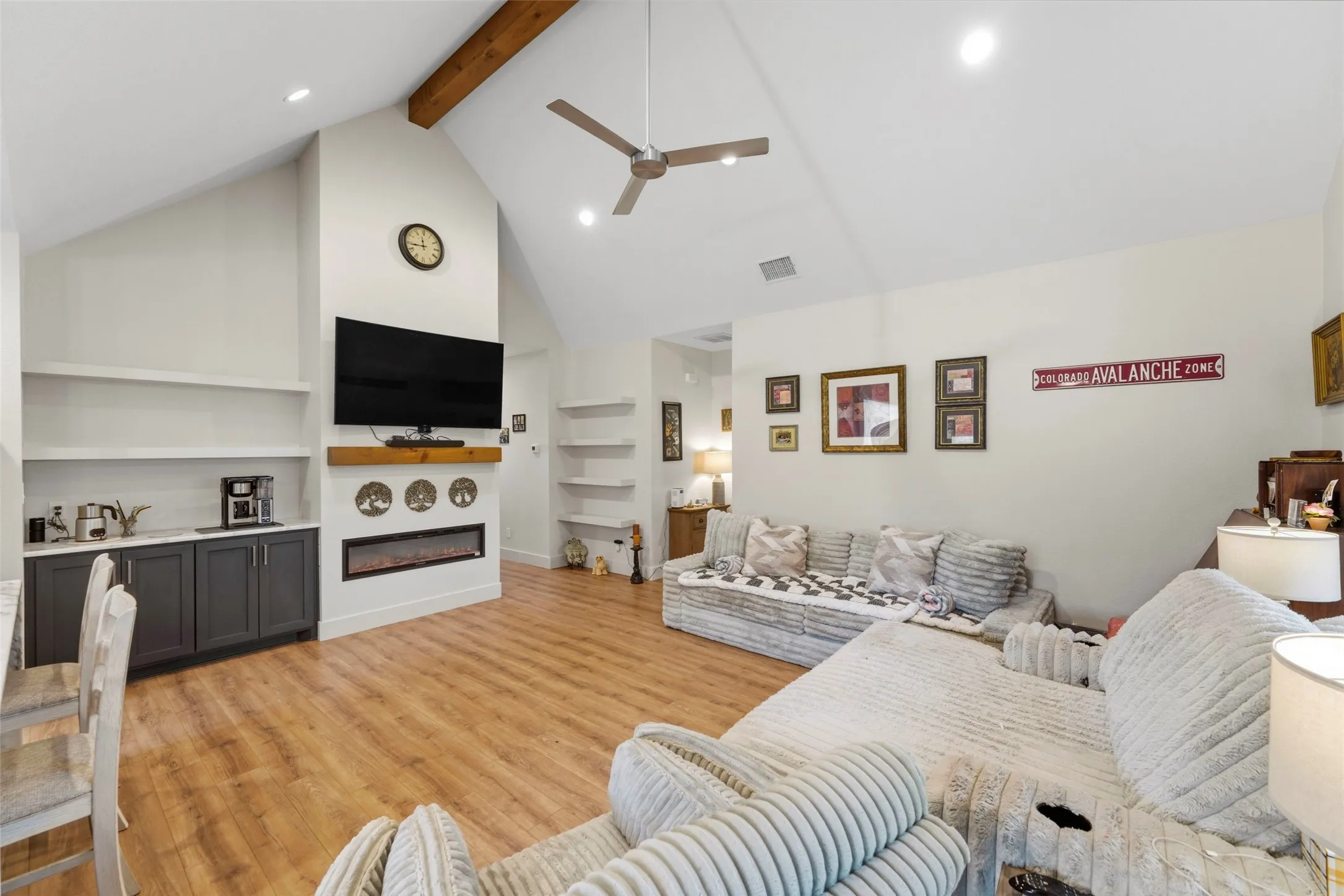 Living area featuring a vaulted ceiling with exposed wood beam, wood-finish flooring, built-in shelving, a modern electric fireplace, and a built-in wet bar with dark cabinetry
