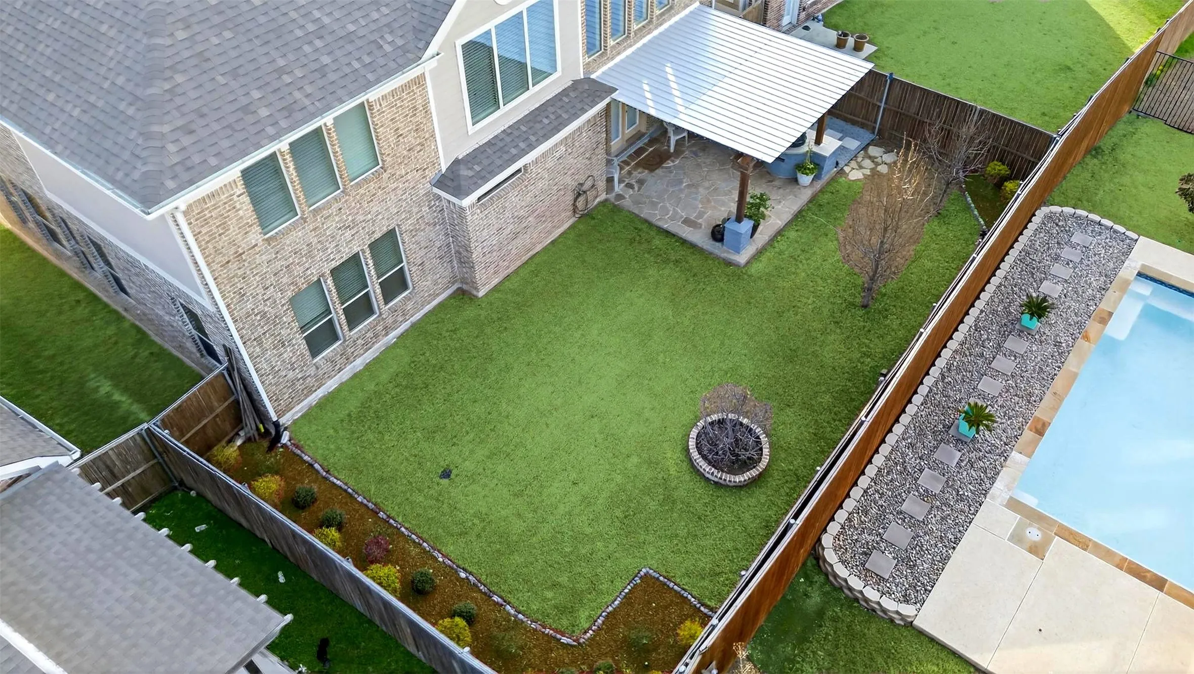 Expansive backyard featuring a swimming pool with concrete surround, artificial turf lawn, built-in fire pit, and a covered patio with stone pavers