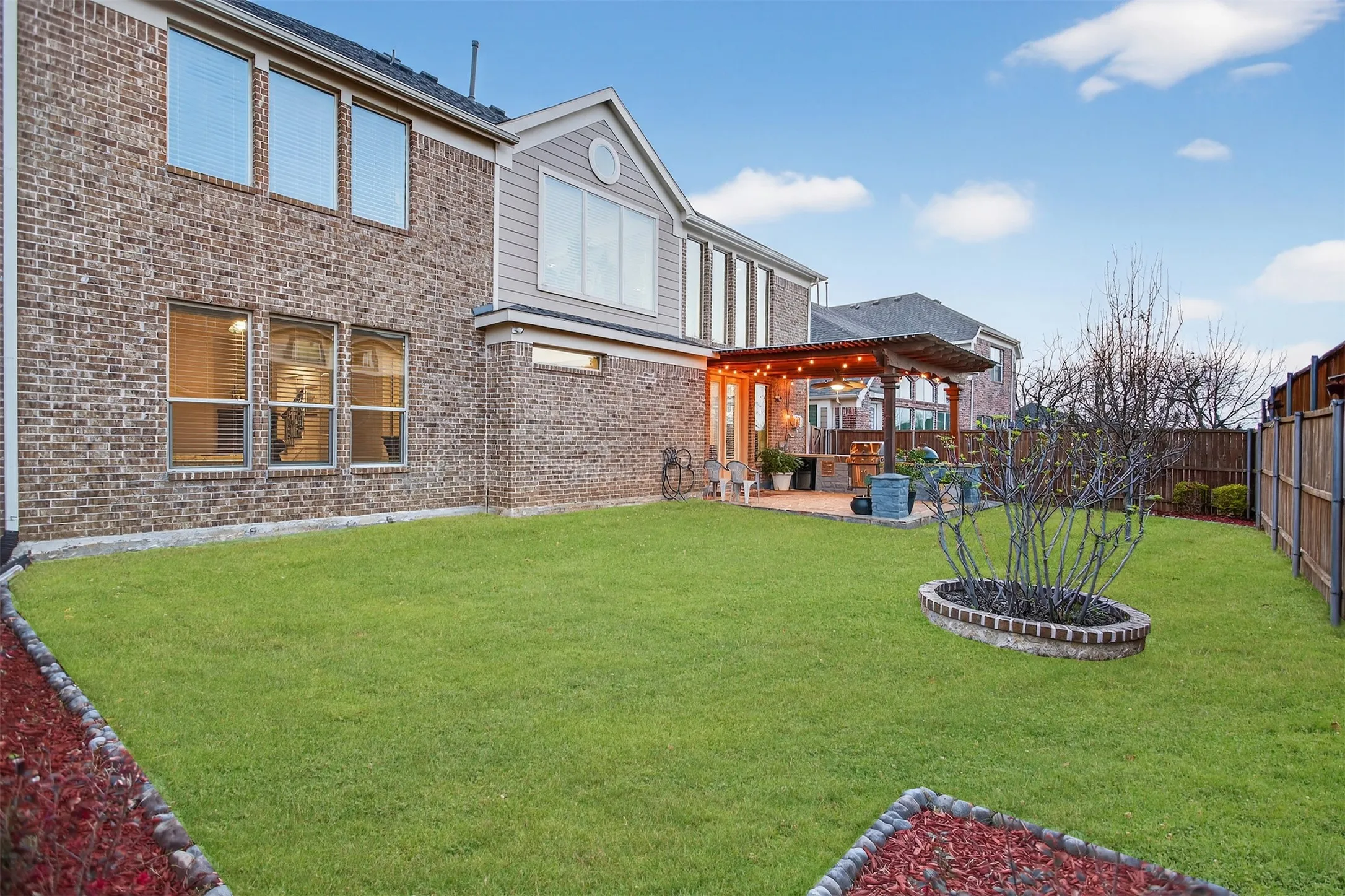 Brick exterior with multiple windows, covered patio with string lighting, paved patio area, and a fenced backyard