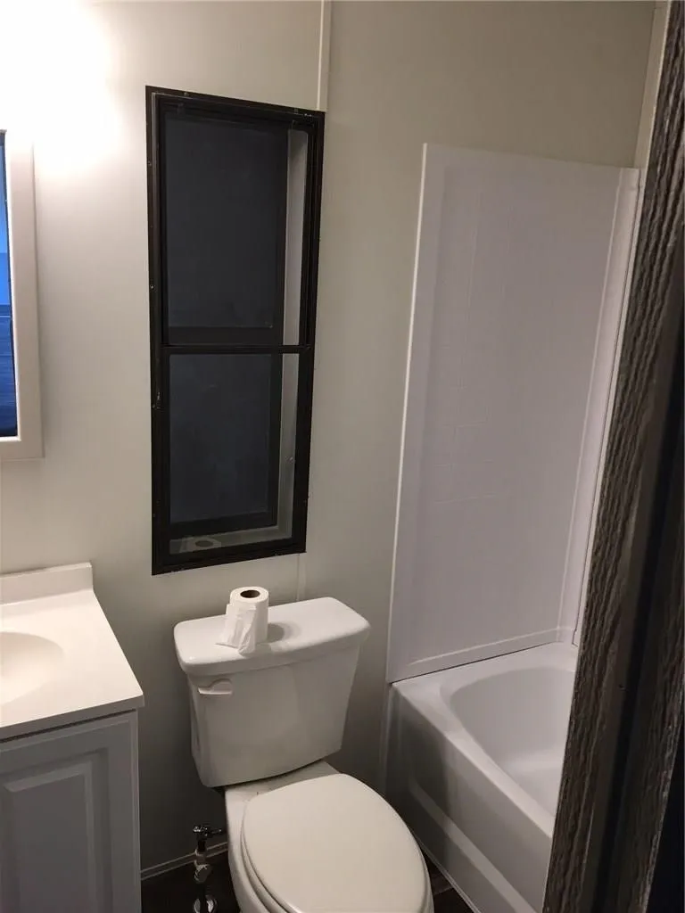 White vanity with integrated sink, white toilet, white bathtub with wall surround, and dark-framed vertical window