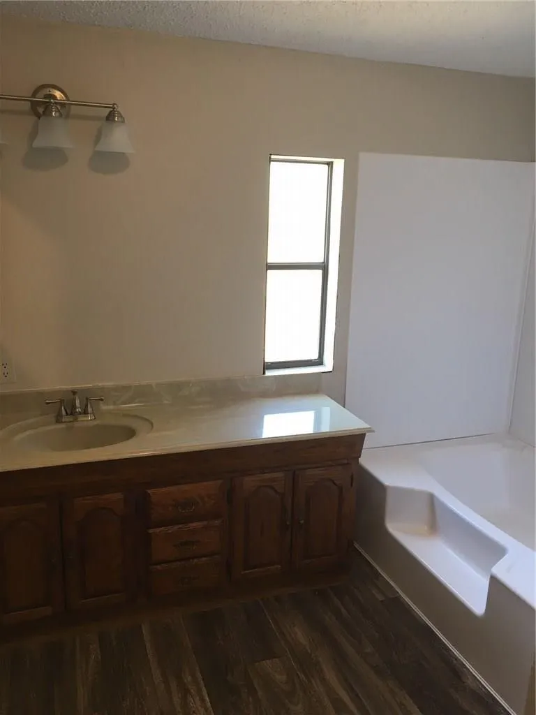 Wood-finish flooring, wood vanity with integrated sink, light countertop, three-light vanity fixture, and a built-in tub