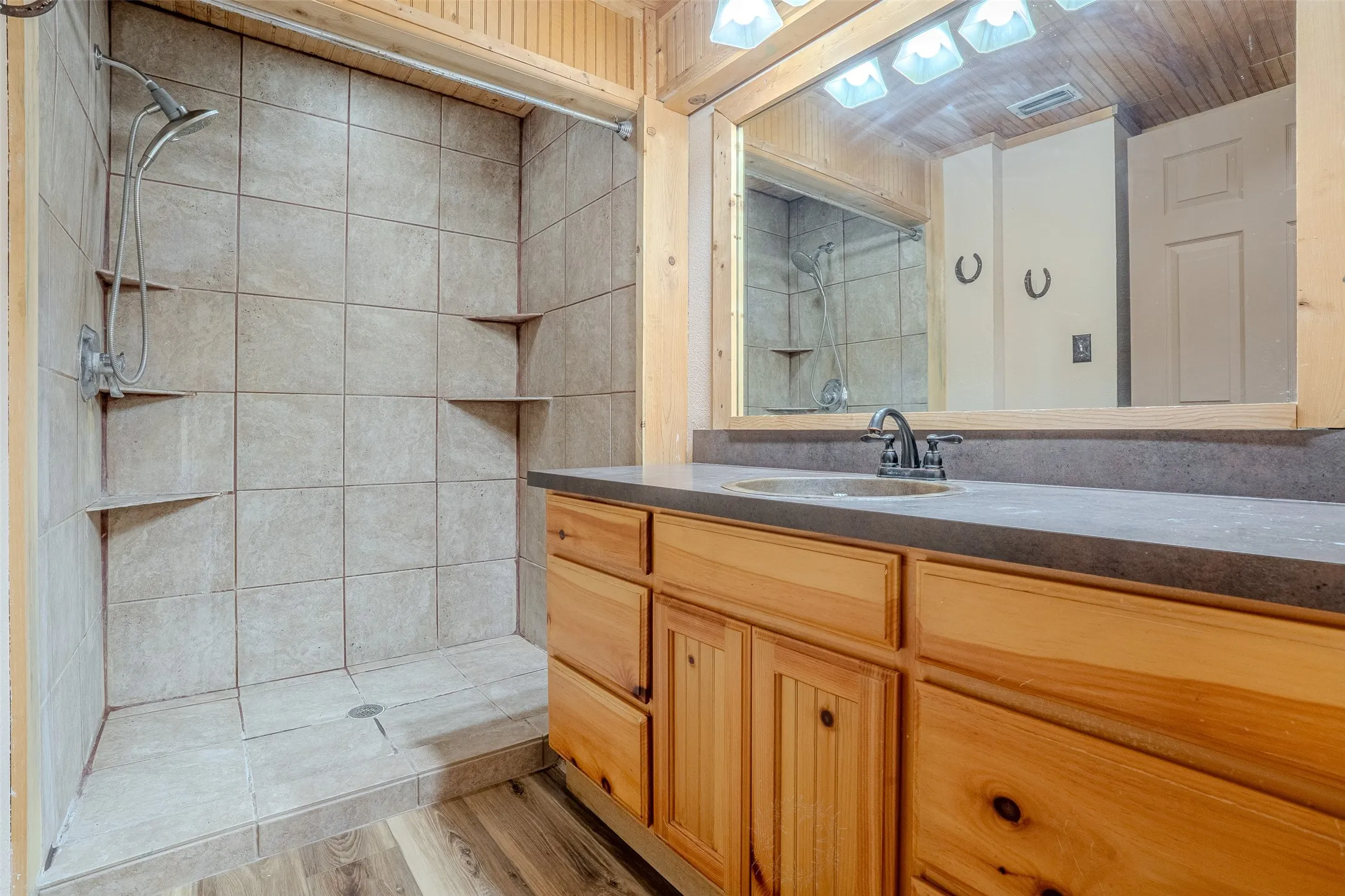 Wood-finish flooring, wood cabinetry with ample storage, dark countertop, and a tiled shower with integrated shelving