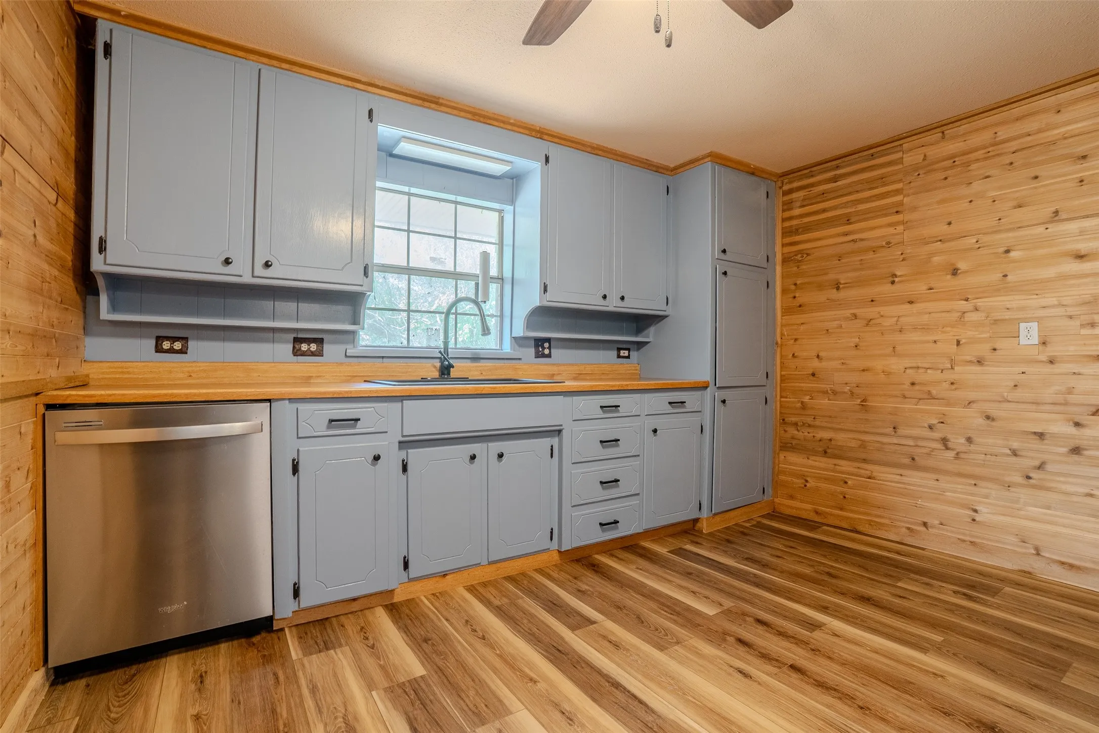 Kitchen featuring wood-finish flooring, light gray cabinetry, natural wood-paneled walls, and a stainless-steel dishwasher