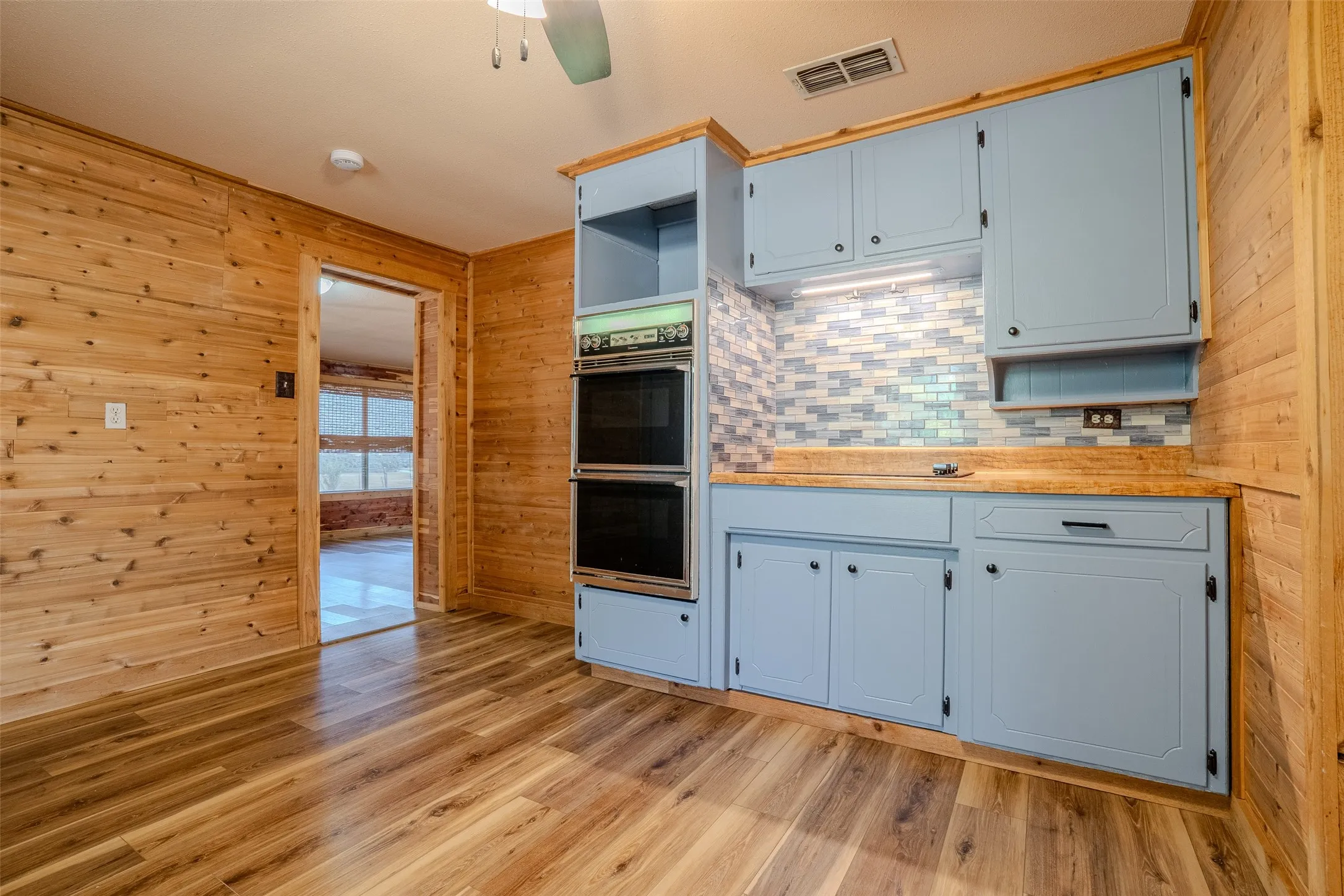 Wood-finish flooring, wood paneling, light blue cabinetry, mosaic tile backsplash, and built-in double ovens