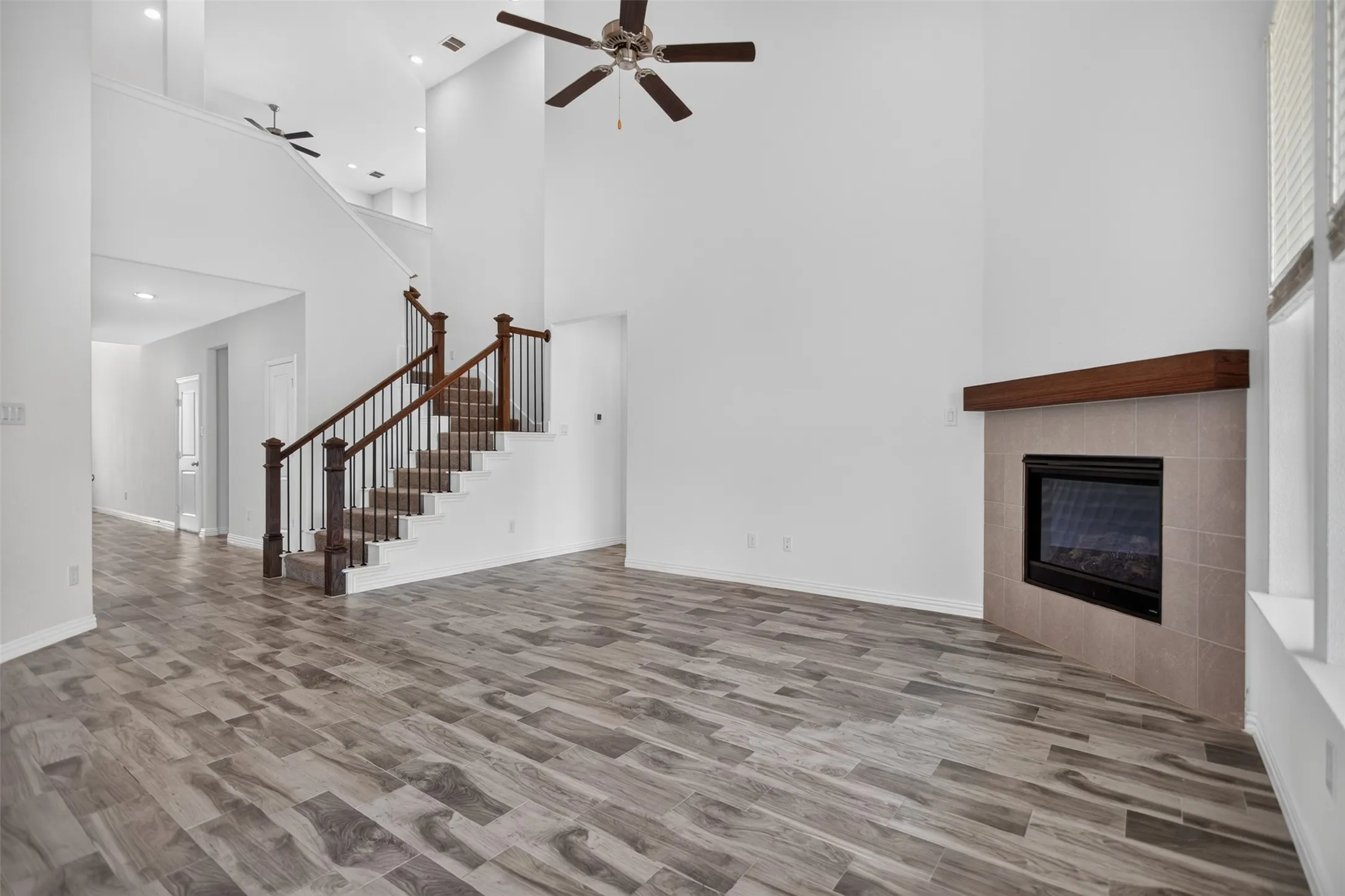 Two-story ceiling, wood-finish flooring, staircase with wood banister and metal spindles, recessed lighting, and a fireplace with tile surround and wood mantle