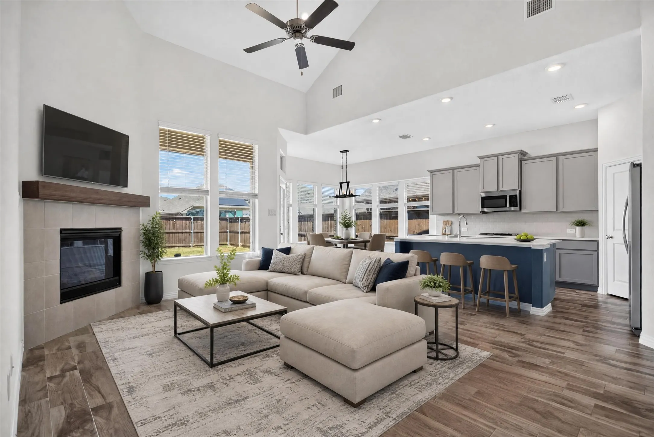Vaulted ceiling with ceiling fan, tile-surround fireplace with wood-finish mantel, wood-finish flooring, kitchen island with seating, and stainless steel appliances