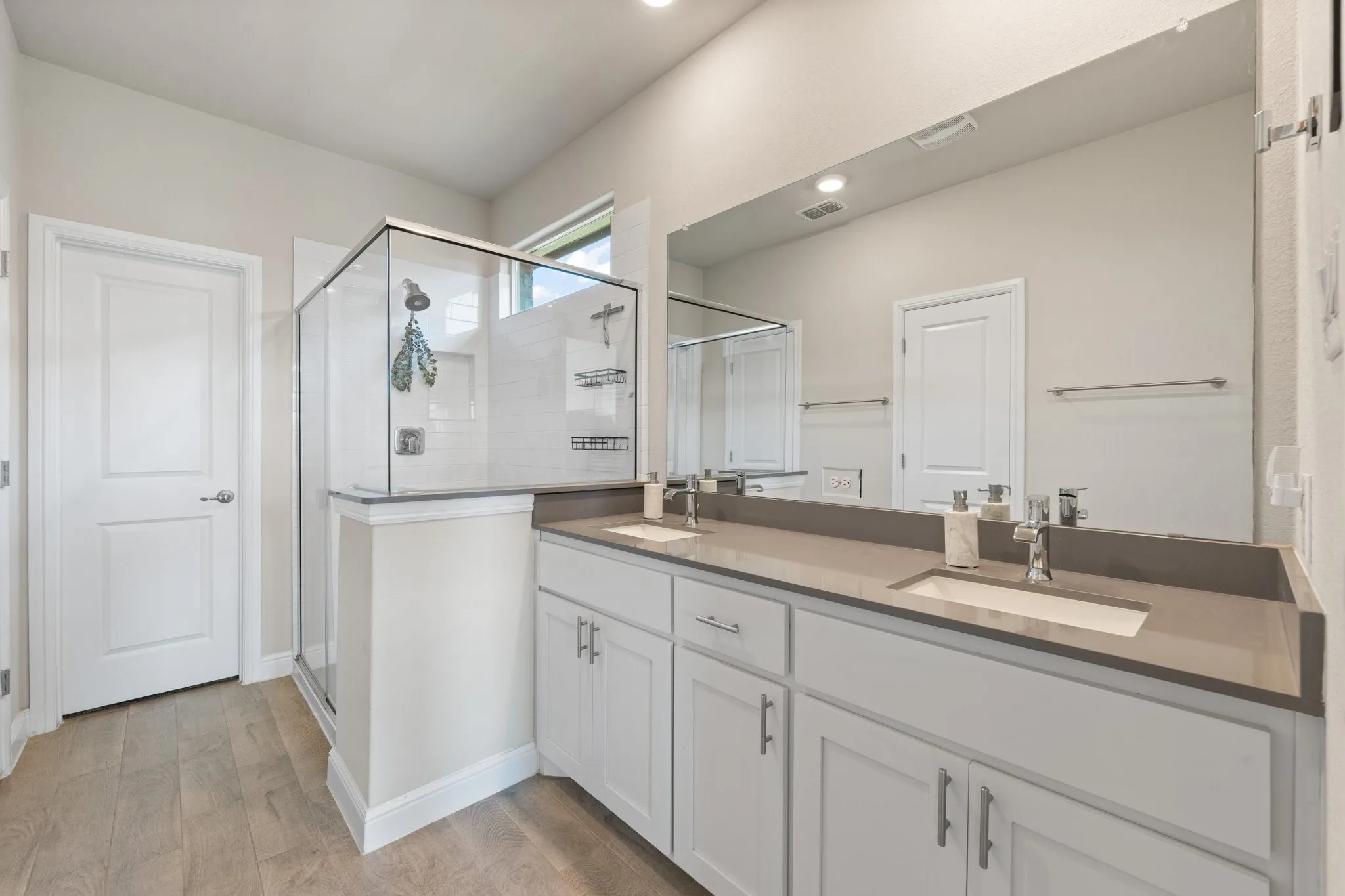 Double vanity with gray countertops, wood-finish flooring, glass-enclosed shower, and a large wall-mounted mirror