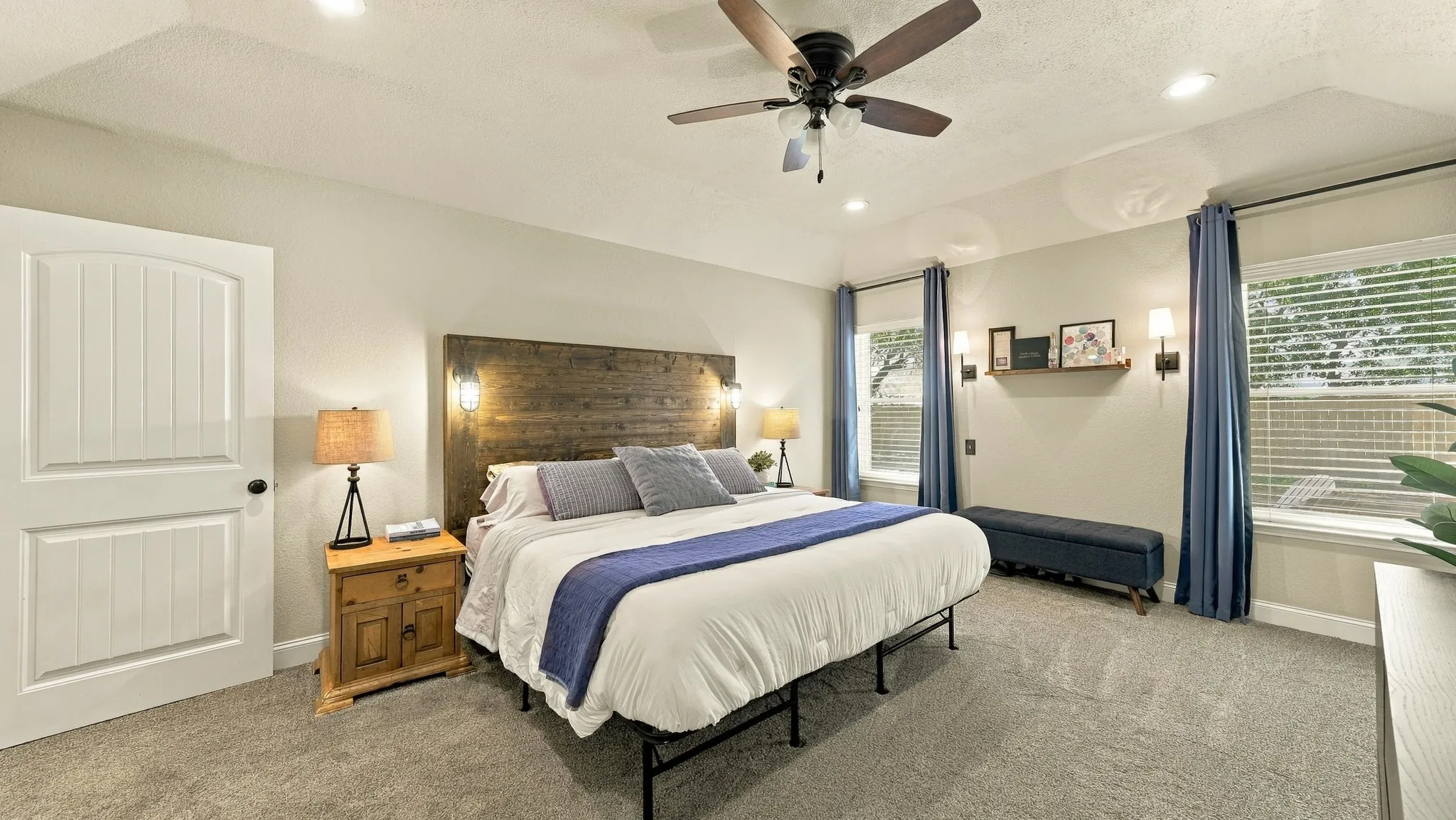 Recessed lighting, ceiling fan, wood-finish headboard with integrated sconces, two windows with blinds, and white paneled door