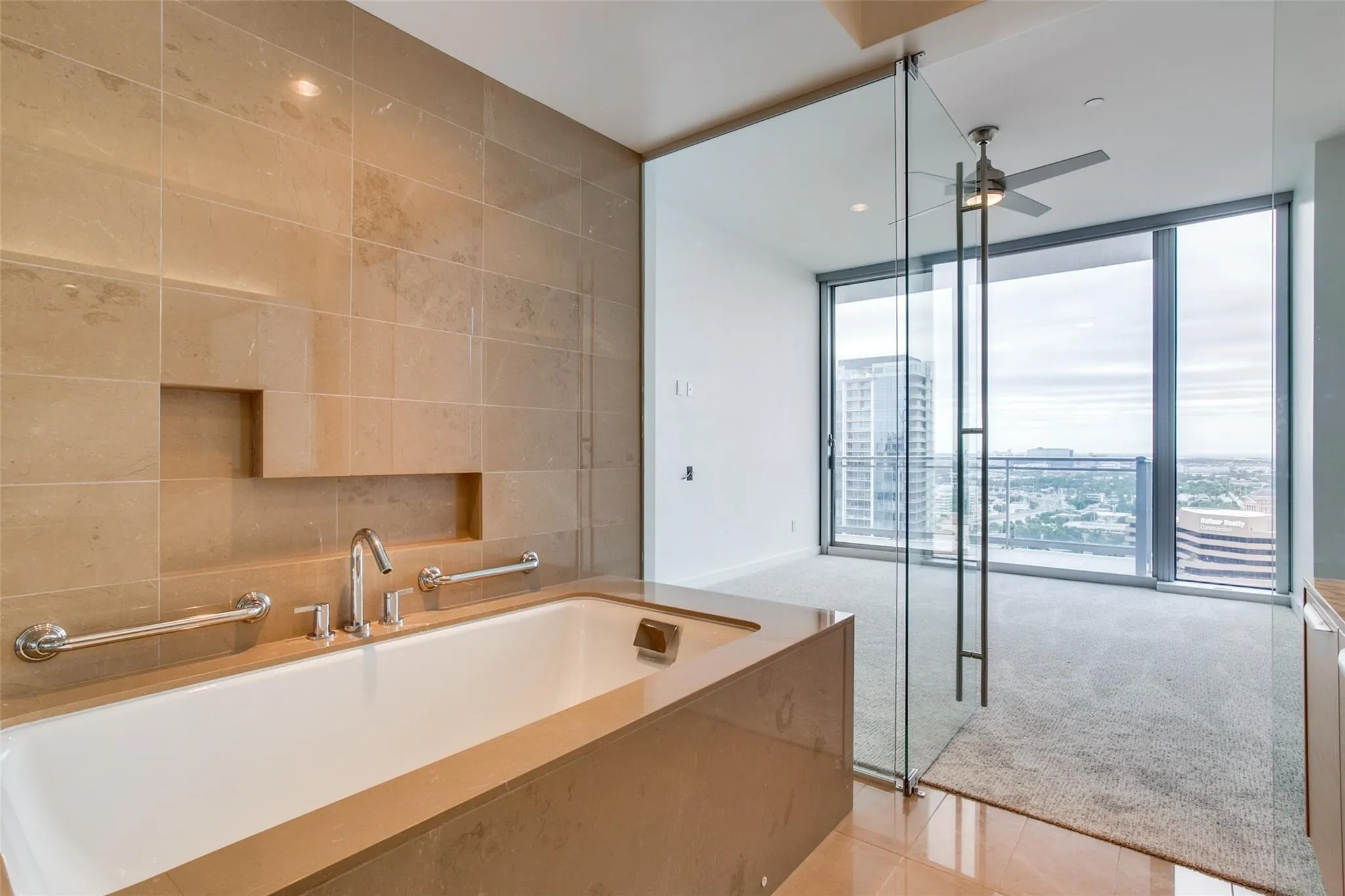 Soaking tub with integrated shelf and chrome fixtures, full-height tile surround, glass partition, floor-to-ceiling windows, and ceiling fan