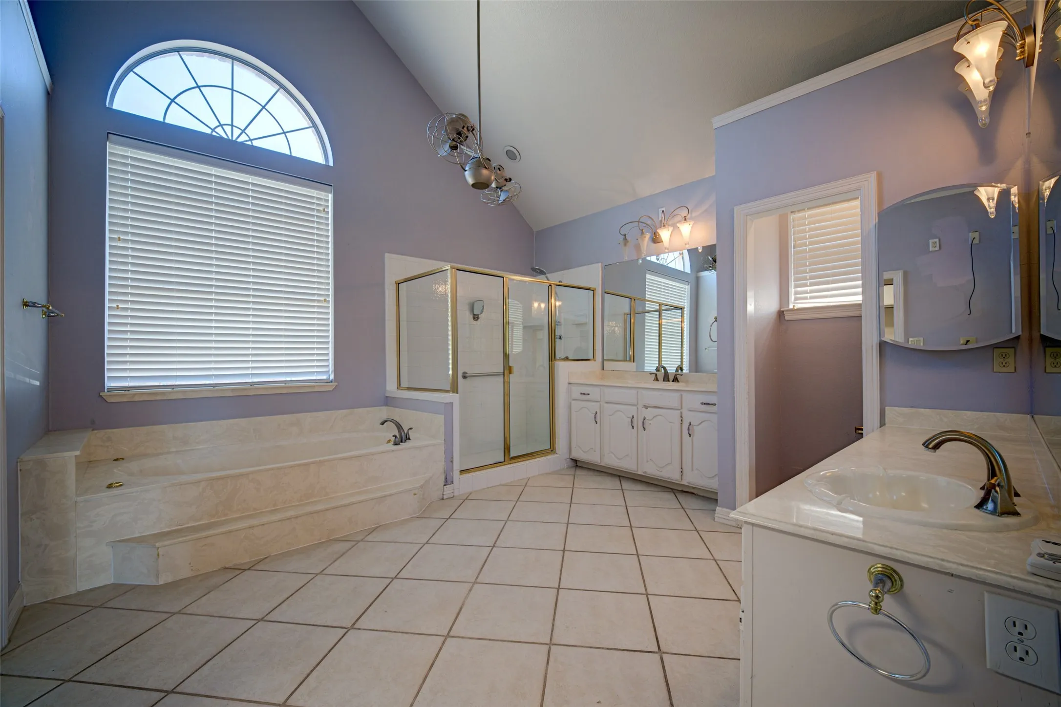 Vaulted ceiling with an arched window, built-in soaking tub, separate shower enclosure with brass-finish framing, dual vanity with white cabinetry, and large format tile flooring