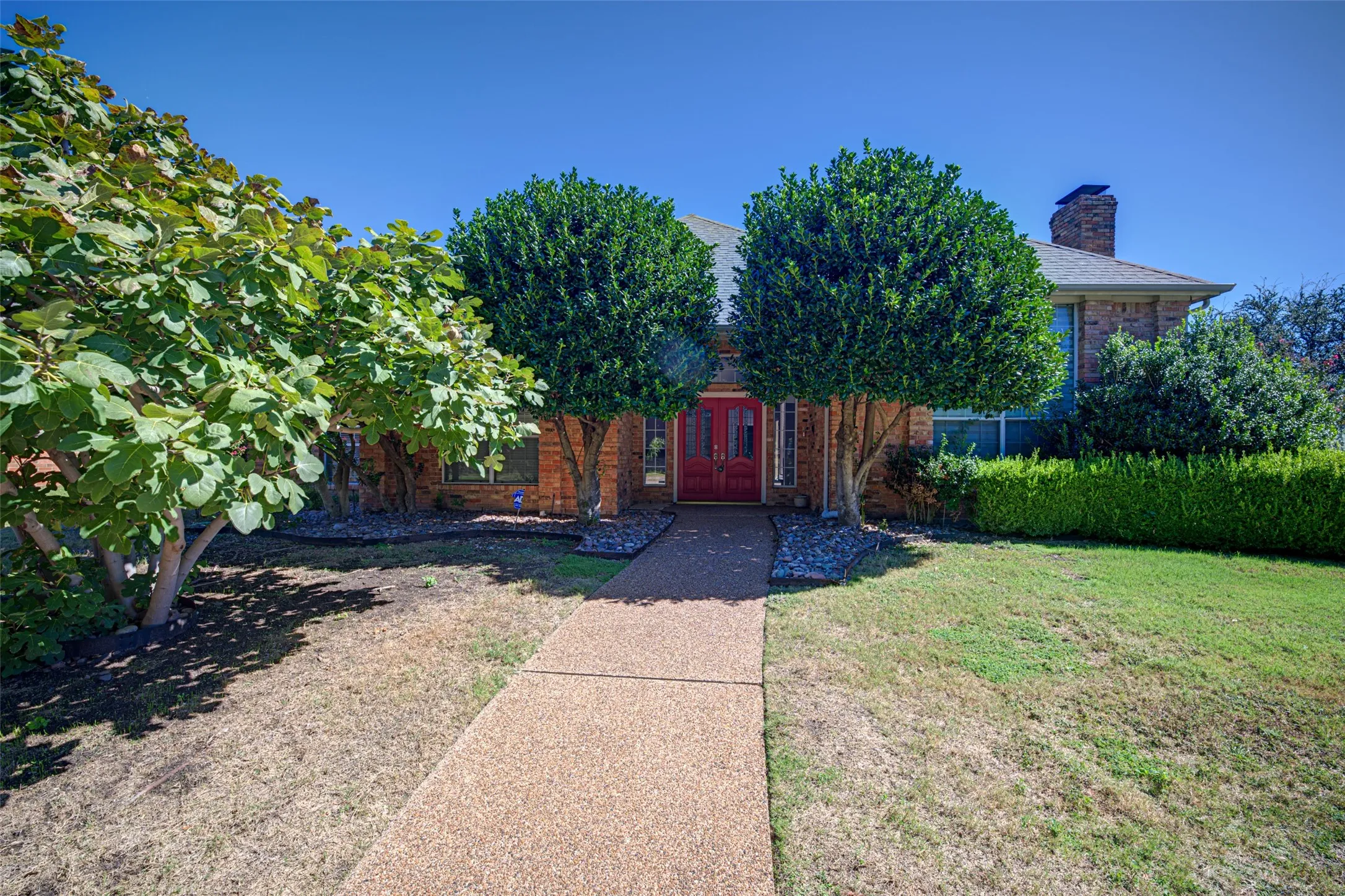 Brick exterior with an exposed aggregate concrete walkway, mature trees, and a red double-door entry