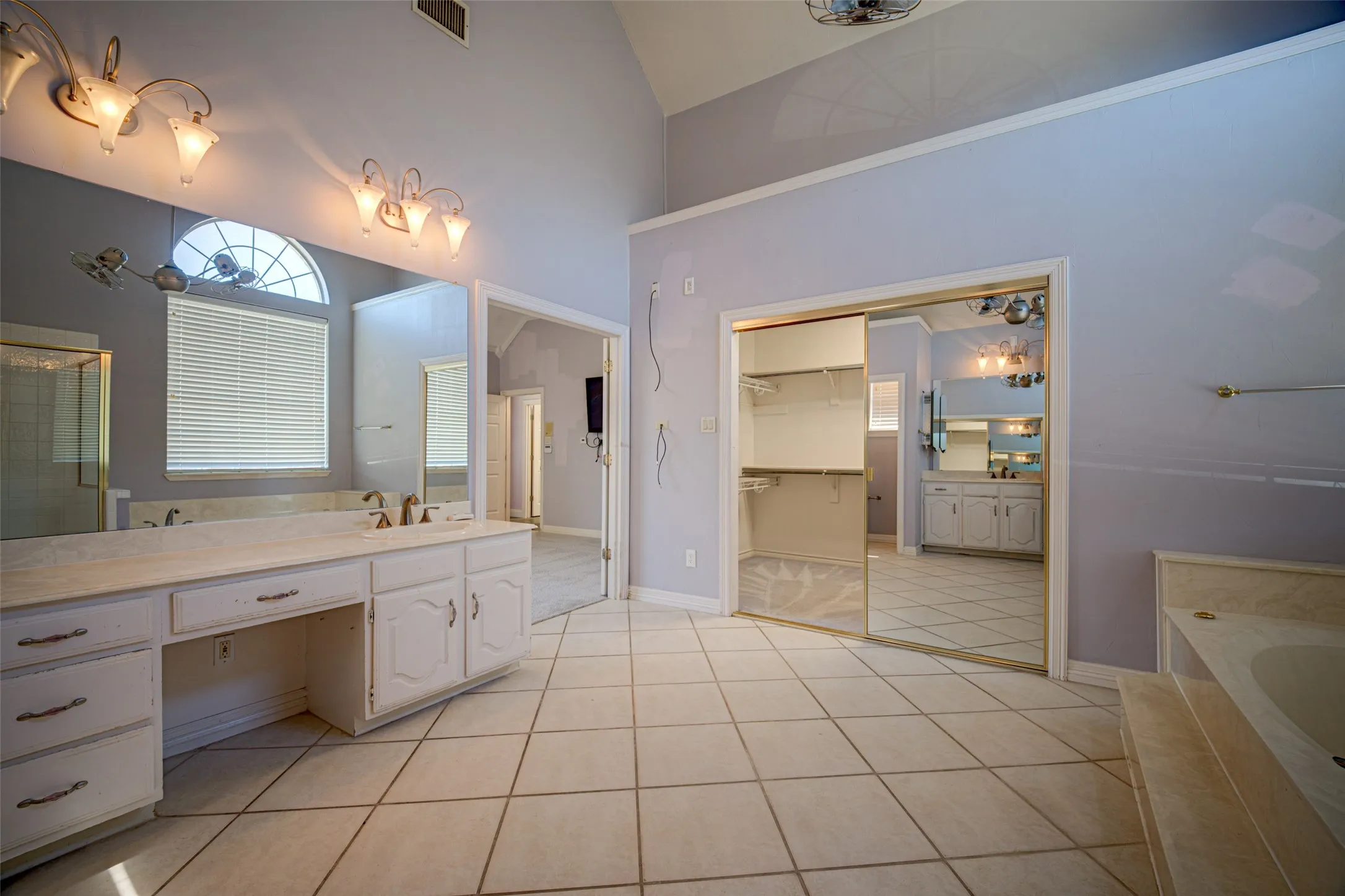 Vaulted ceiling with a ceiling fan, dual vanity with an arched window, tile flooring, and a built-in closet with mirrored sliding doors