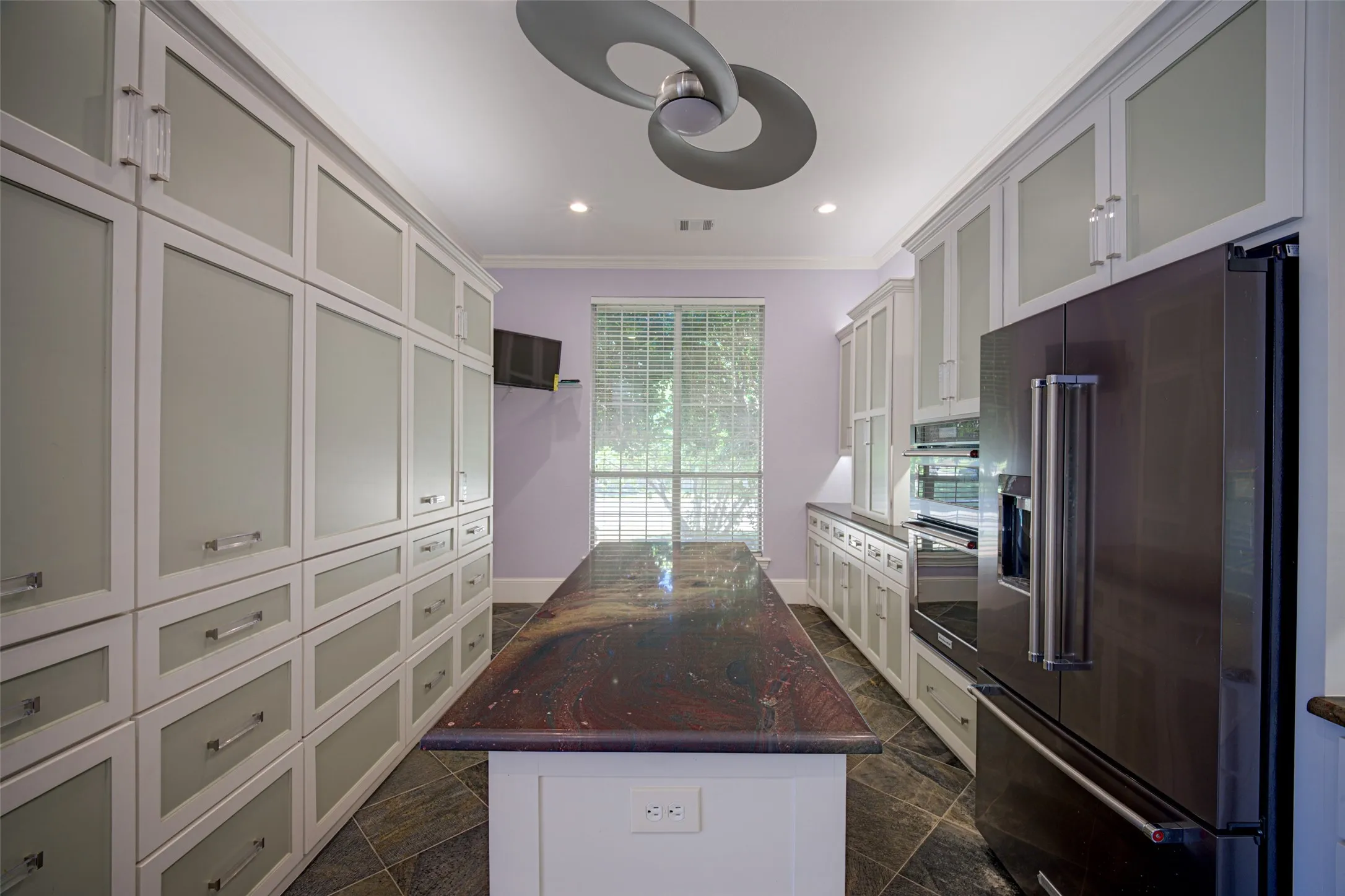 Expansive storage cabinetry with frosted glass panels, an oversized island with a unique countertop, black stainless steel built-in appliances, recessed lighting, and a modern ceiling fan