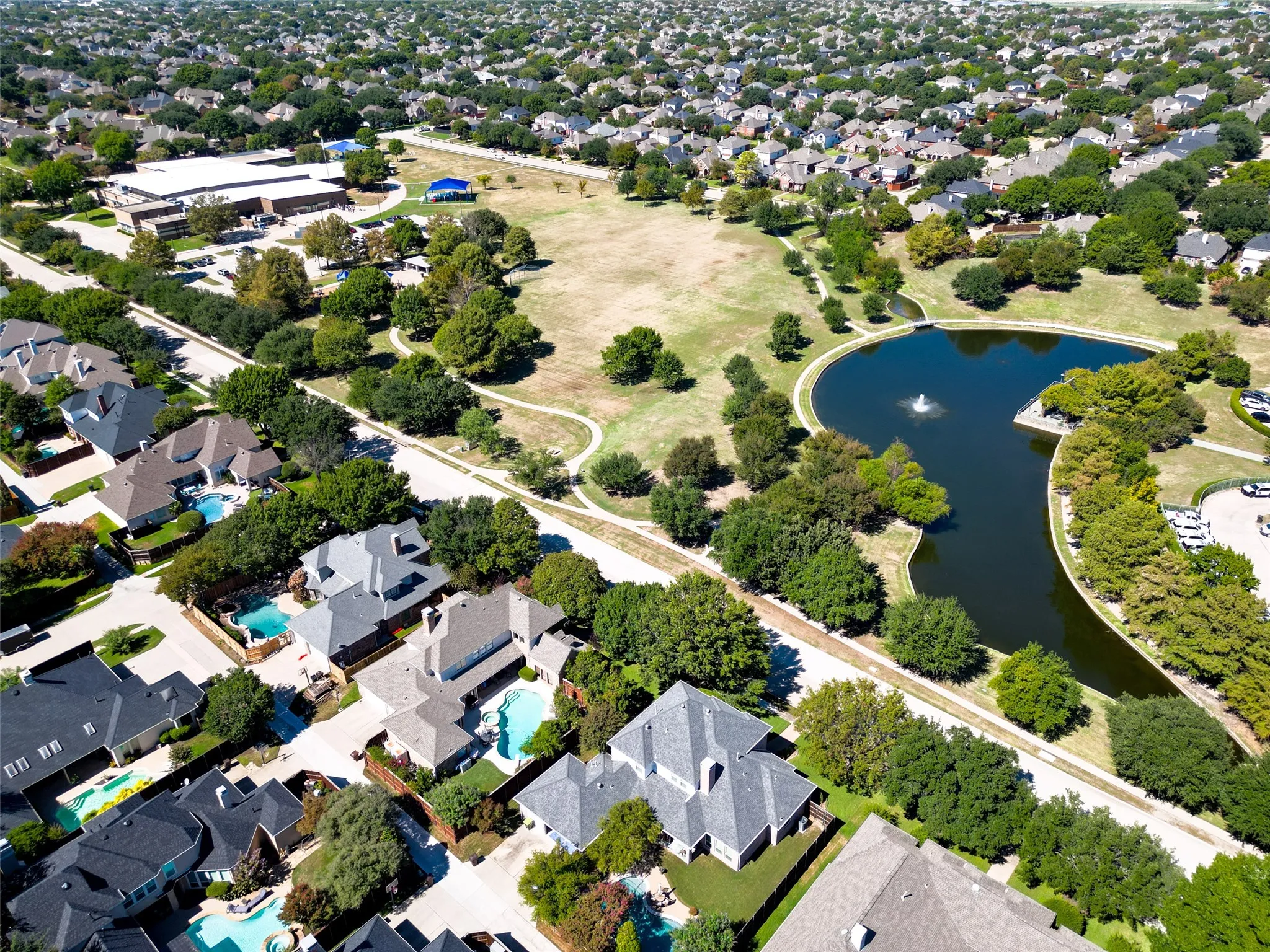 Aerial perspective showcasing a residential community with mature landscaping, a central park, a pond with a fountain, and numerous properties featuring private swimming pools