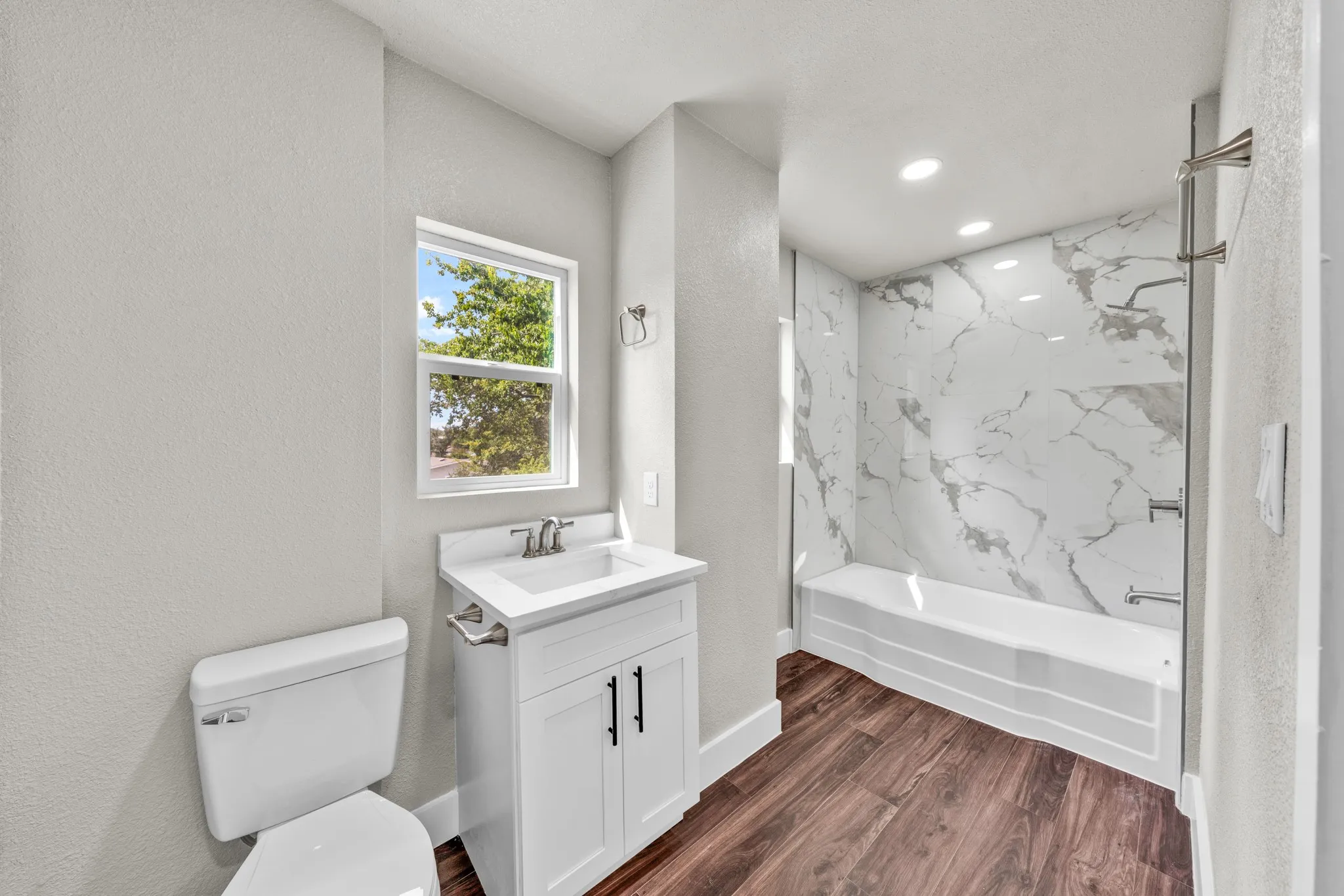Bathroom featuring a white vanity with integrated sink, chrome fixtures, wood-finish flooring, a white toilet, and a bathtub with a marble-patterned surround