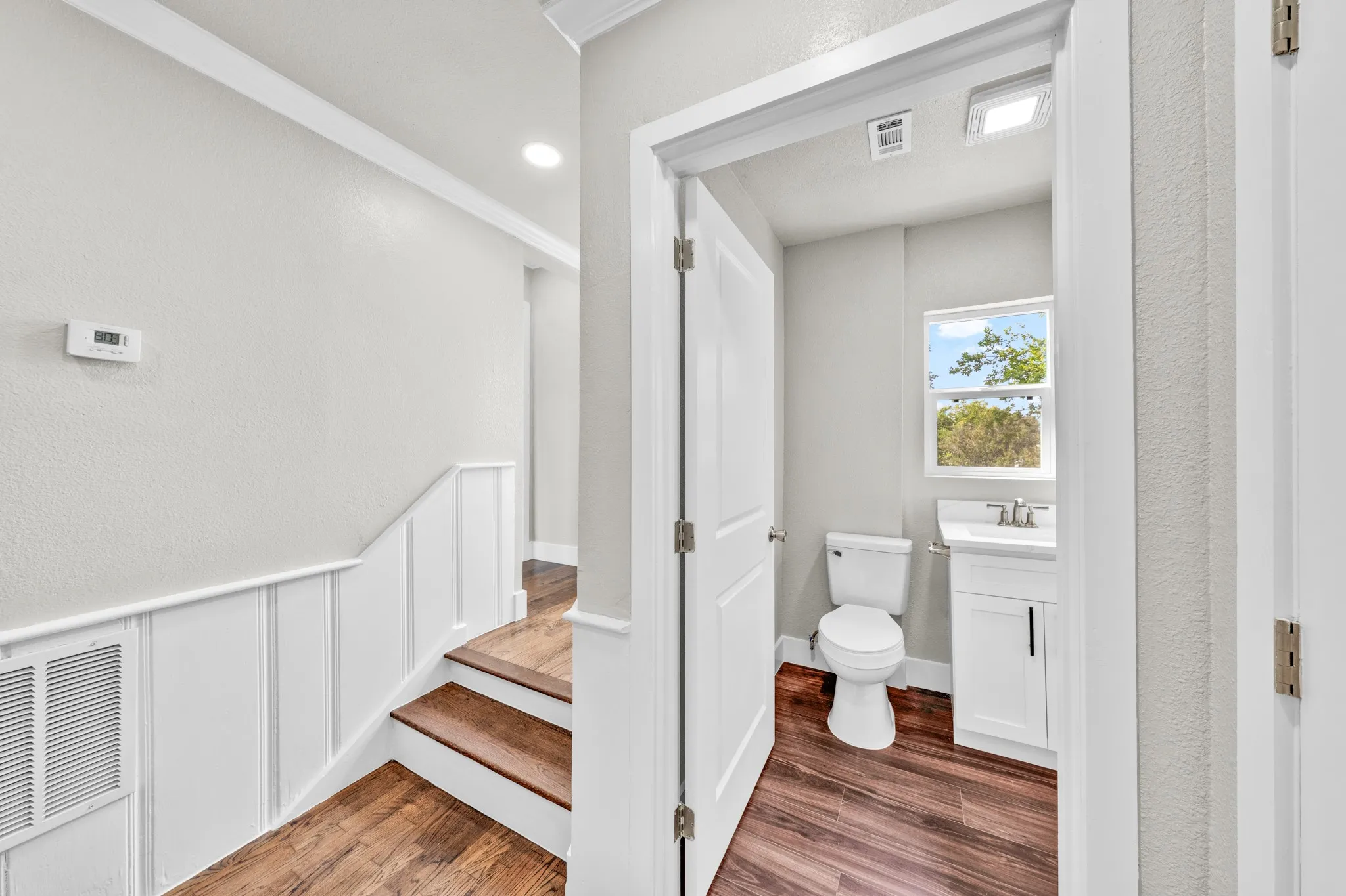 Wood-finish flooring throughout, white wainscoting, recessed lighting, and a bathroom with a window and white vanity