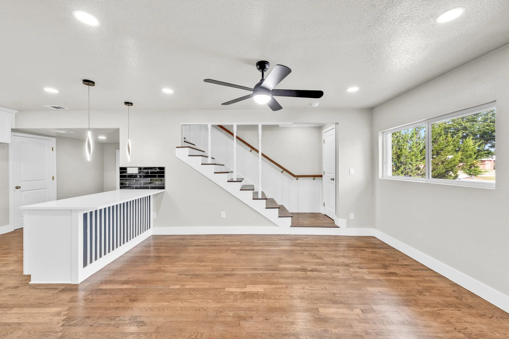Wood-finish flooring throughout, bright white walls, modern ceiling fan, recessed lighting, and a white staircase with wood handrails