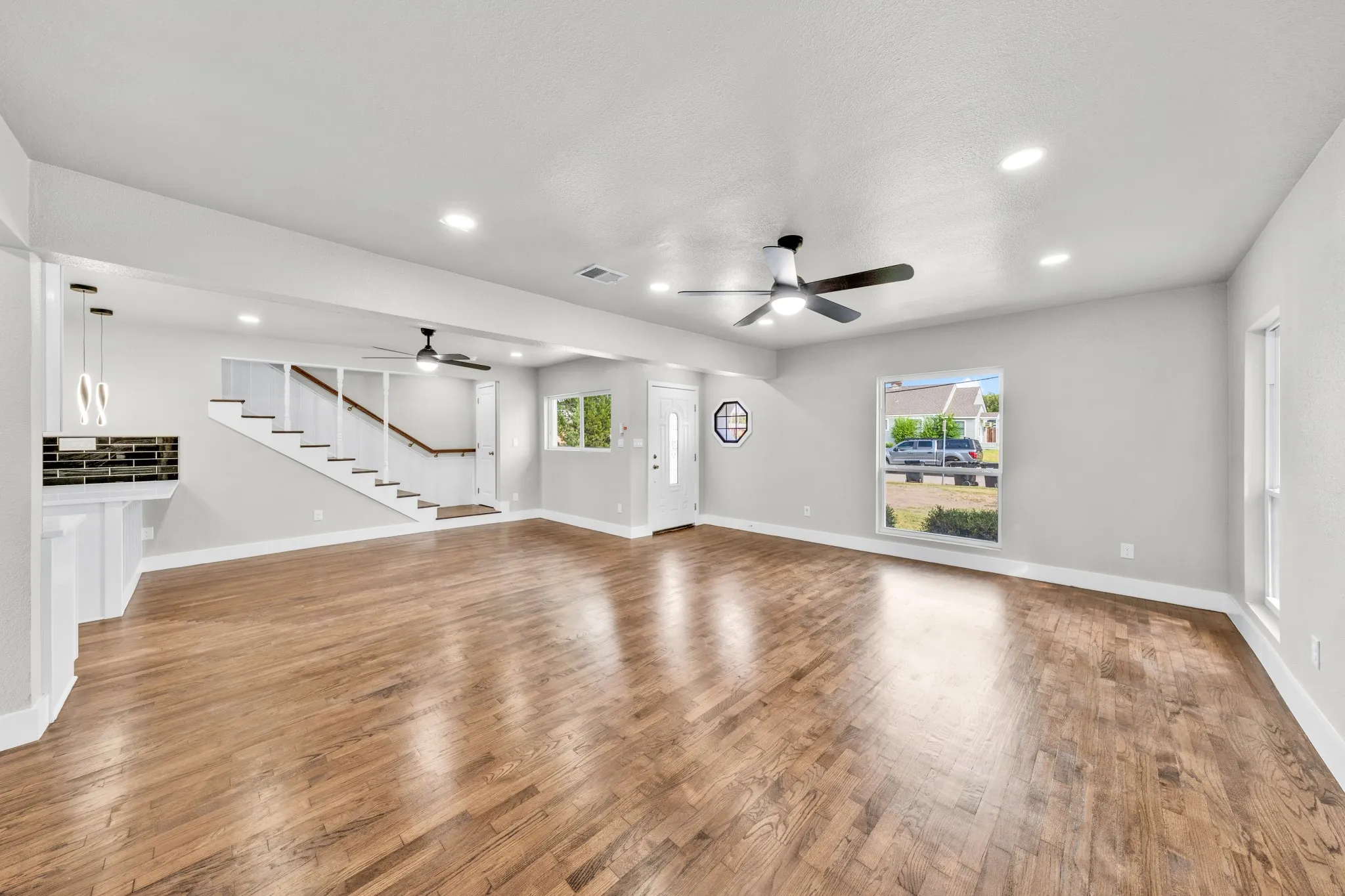 Wood-finish flooring, recessed lighting, ceiling fans, a modern staircase with wood railing, and a tiled accent wall with contemporary pendant lighting